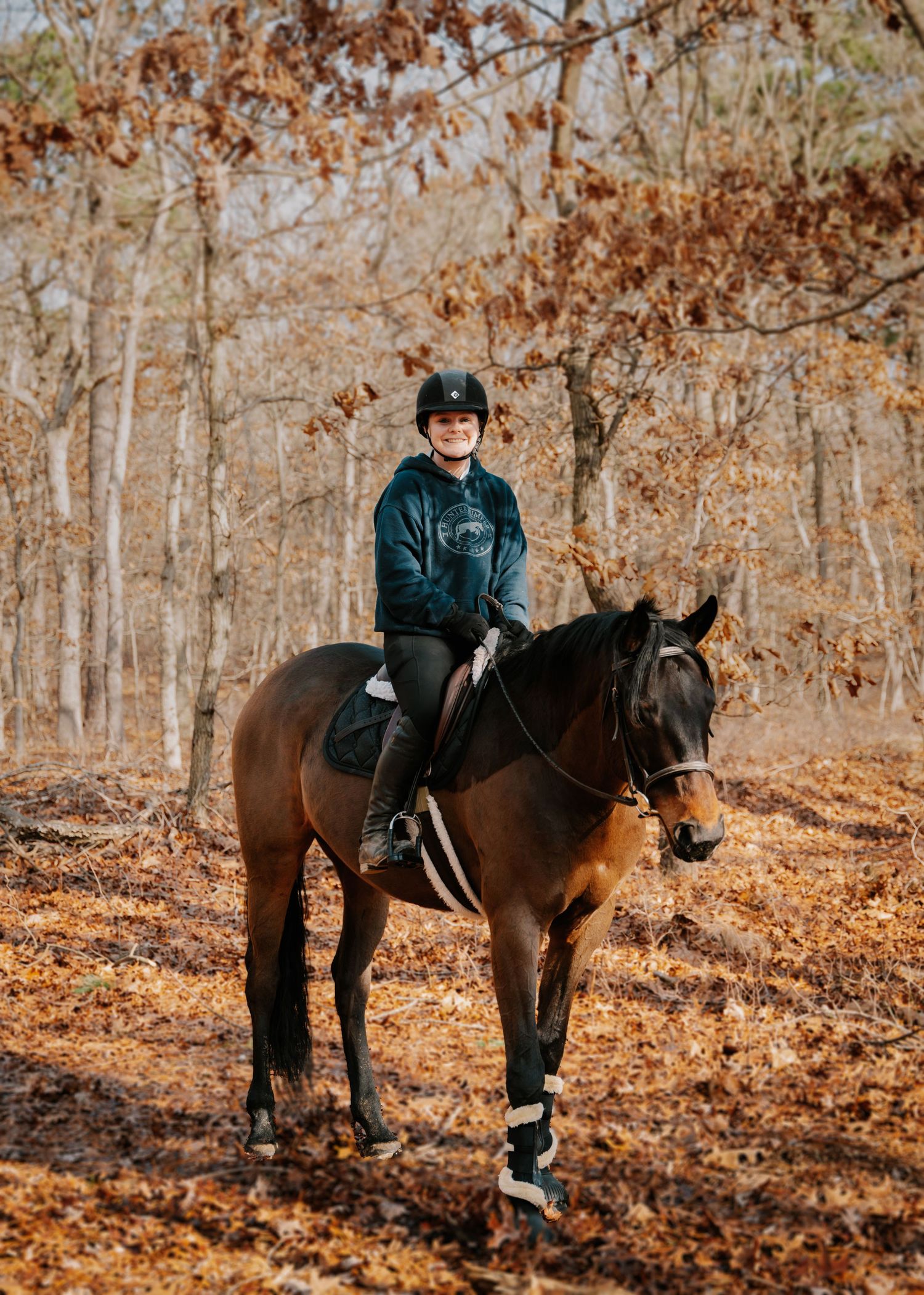A young equestrian in a helmet rides a bay horse through an autumn forest with golden fallen leaves.