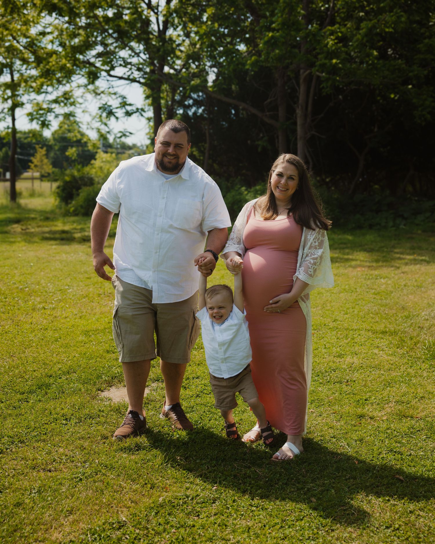 Pregnant woman in pink dress with husband and toddler posing outdoors on green grass with trees in background.