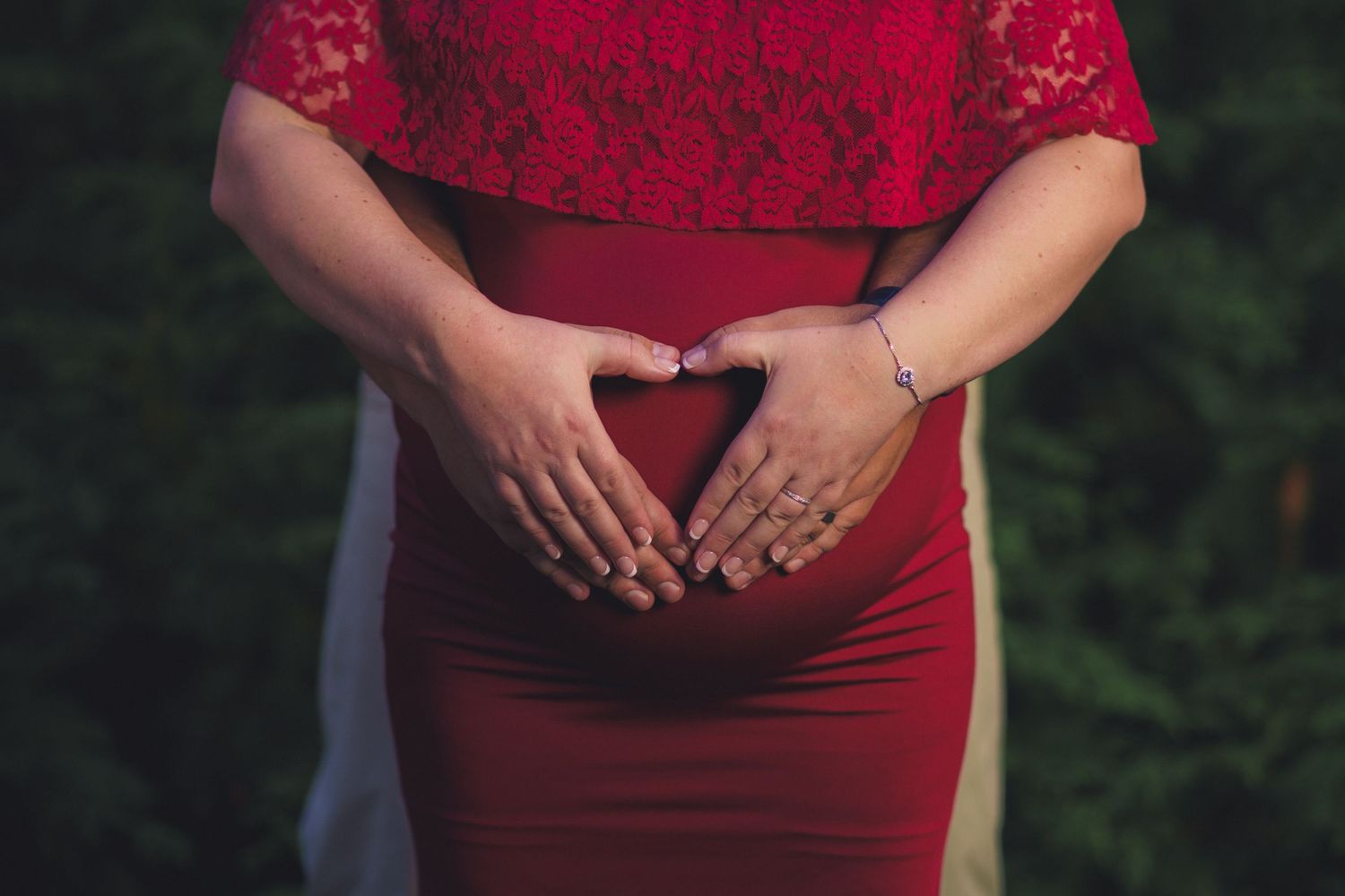Pregnant woman in red lace crop top and skirt forming a heart shape with her hands over her baby bump.