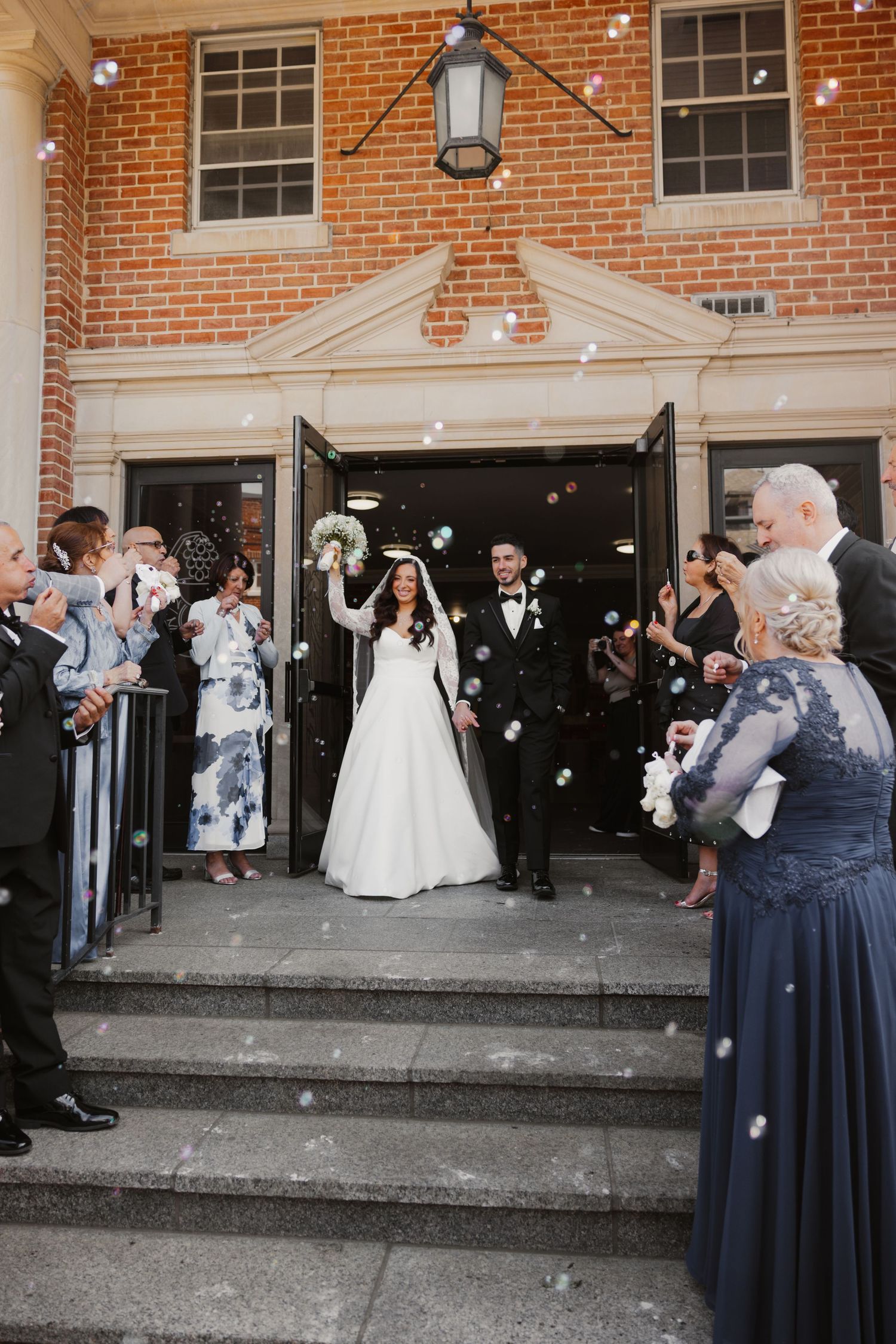 Bride and groom exit brick church surrounded by bubbles and smiling guests in formal attire on wedding day.