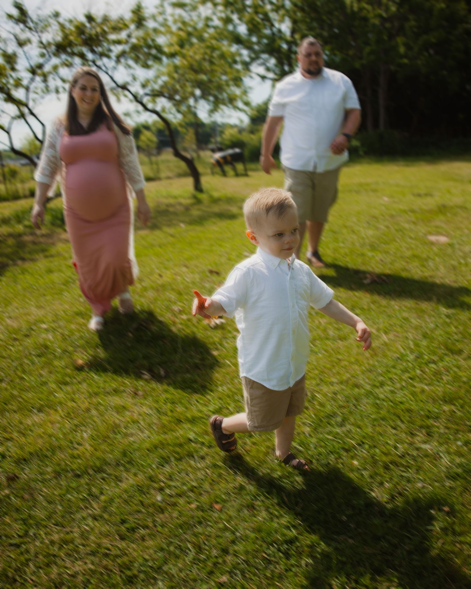 A young boy runs on green grass while his pregnant mother in a pink dress and father walk behind him outdoors.