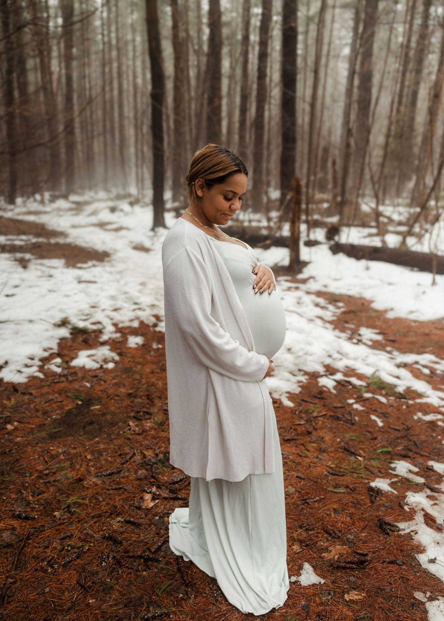 Pregnant woman in white dress and cardigan cradling baby bump in snowy winter forest during maternity photo session.