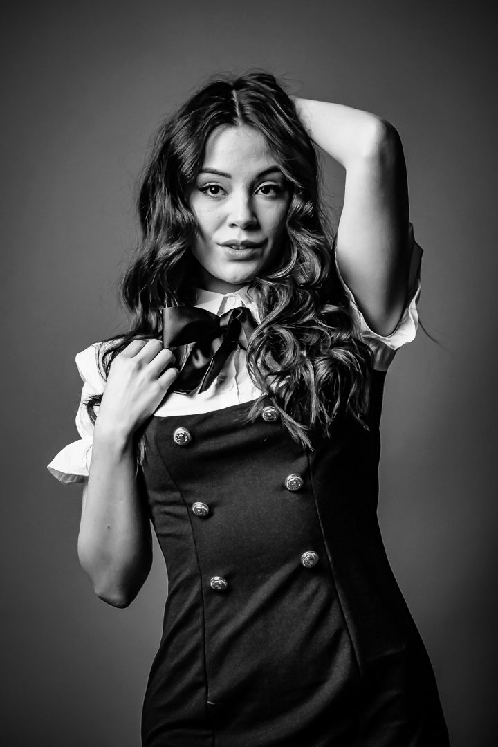 Black and white portrait of a young woman in a dark pinafore dress with bow tie posing with one arm raised.