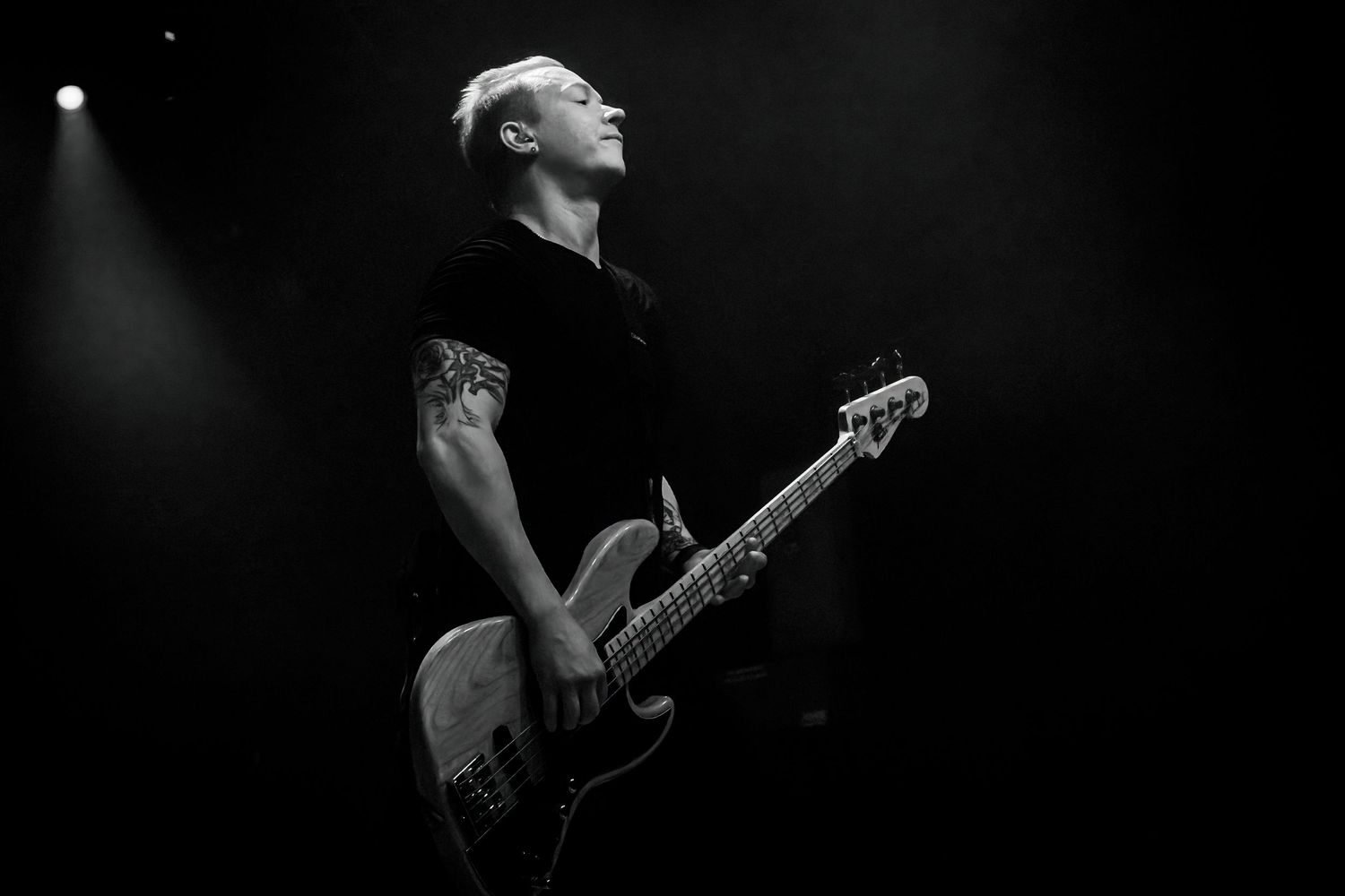 Black and white photo of a bassist playing guitar on stage, head tilted back under a single spotlight.