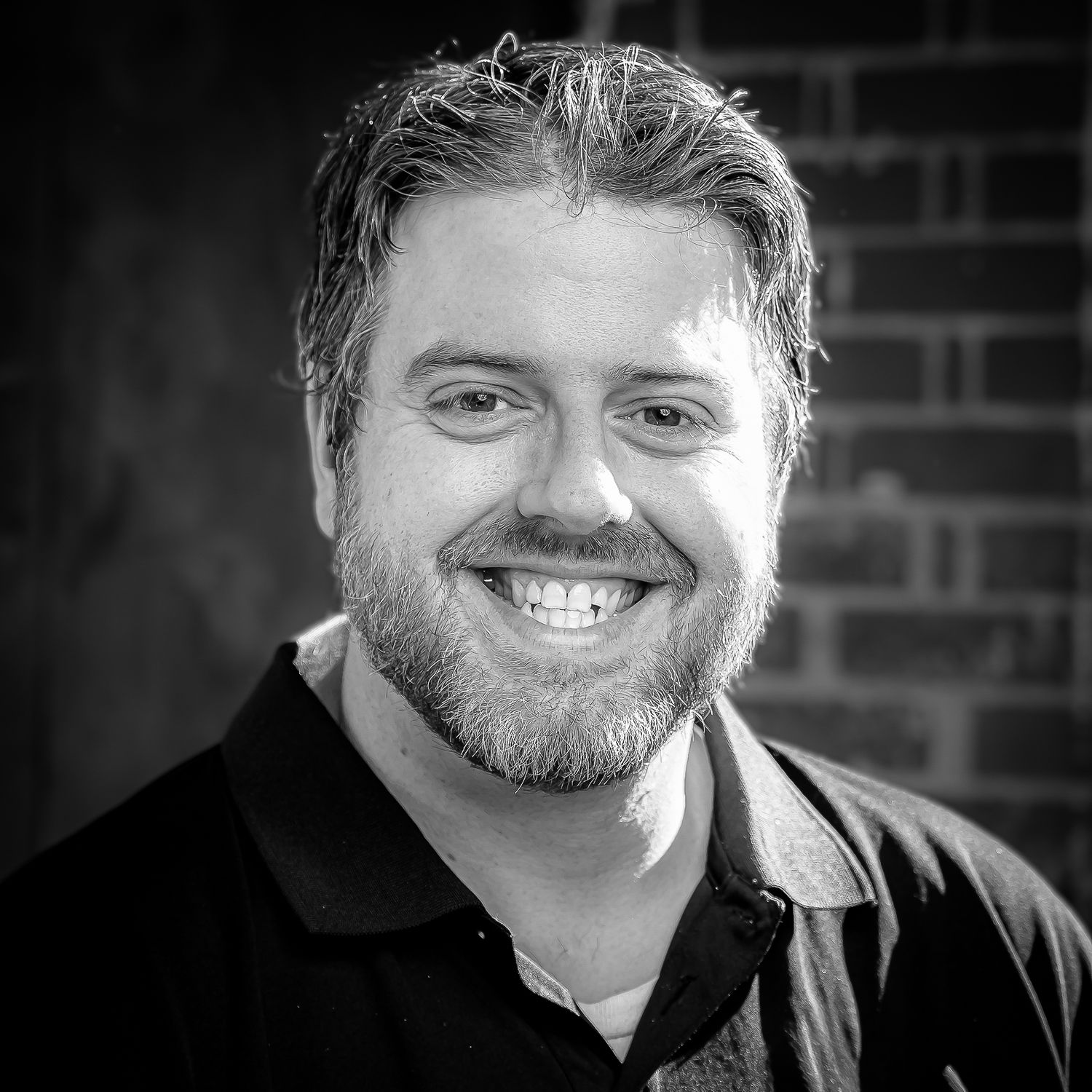 Smiling bearded man in black shirt poses outdoors against brick wall in black and white portrait photo. William Austin Bruce owner WAB Photo