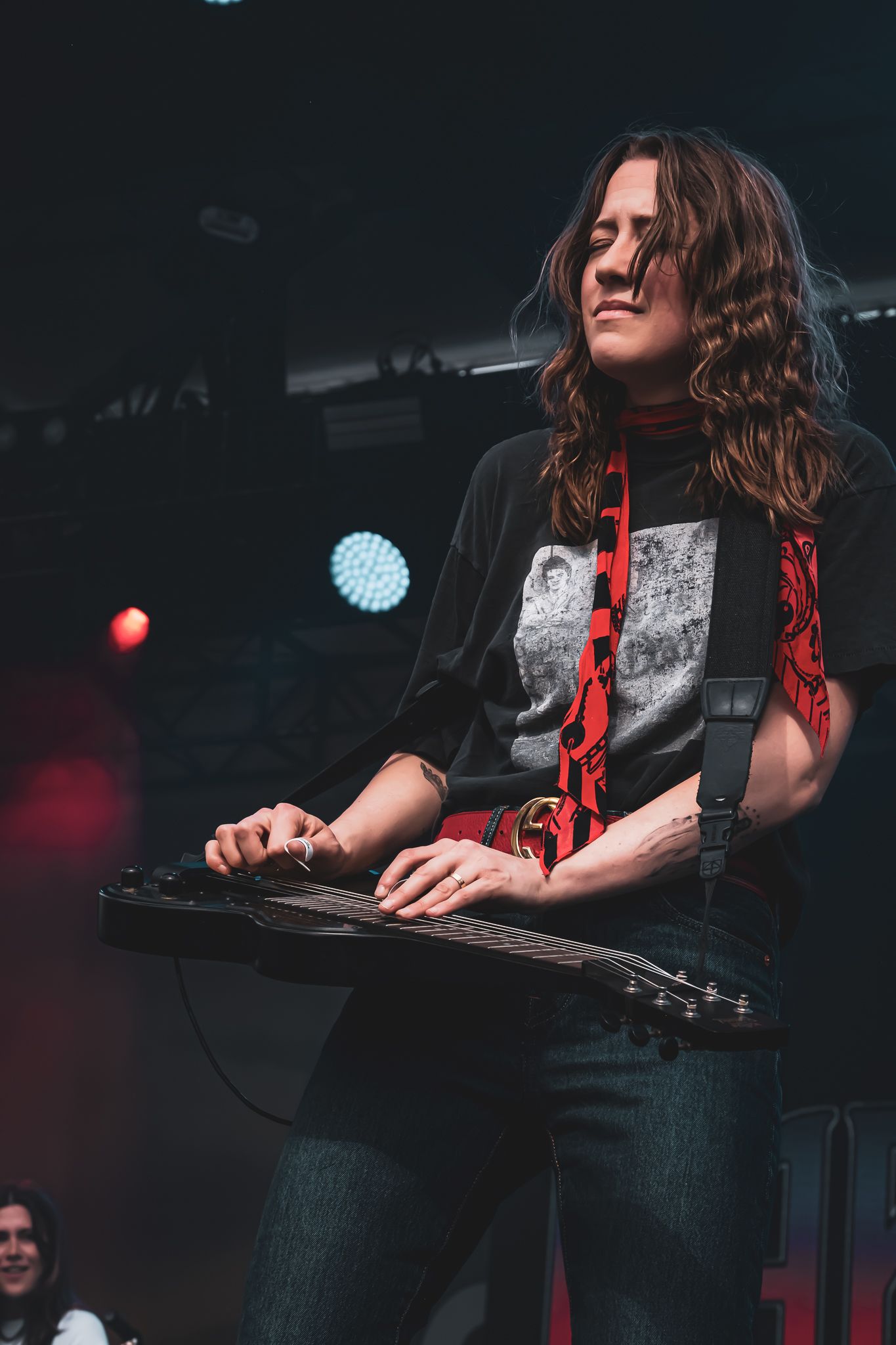 Female musician playing lap steel guitar on stage wearing black graphic tee with red strap.