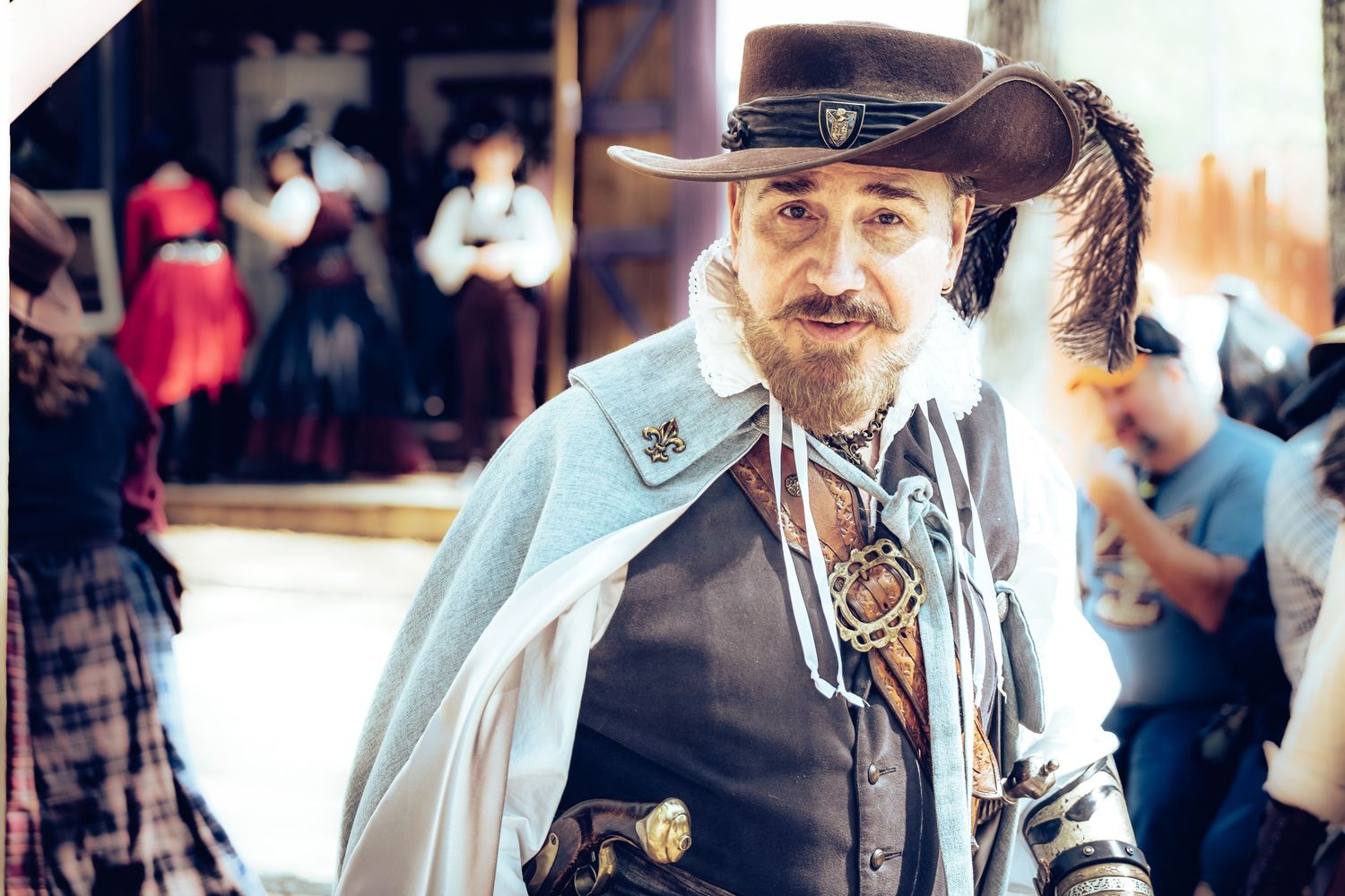 Man in musketeer costume with feathered hat and cape smiles at a Renaissance festival.