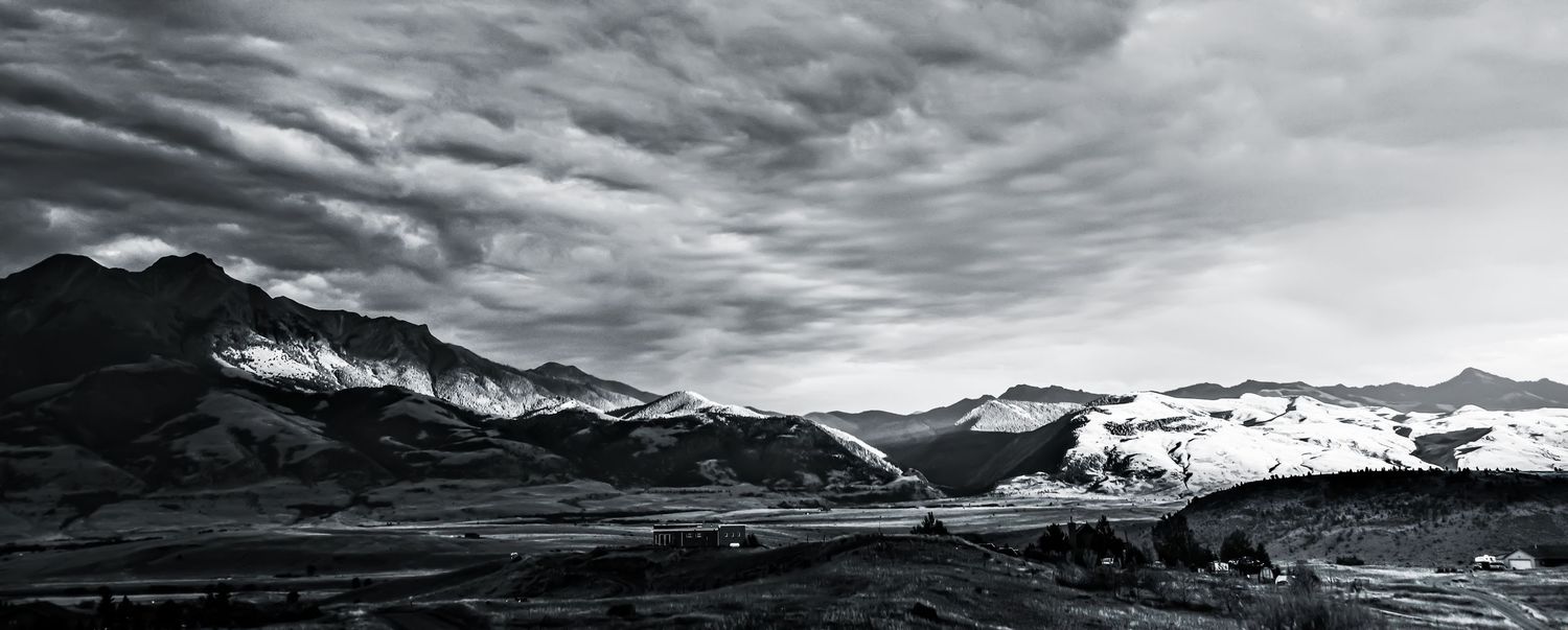 Black and white panoramic landscape of snow-capped mountains under dramatic cloudy skies.