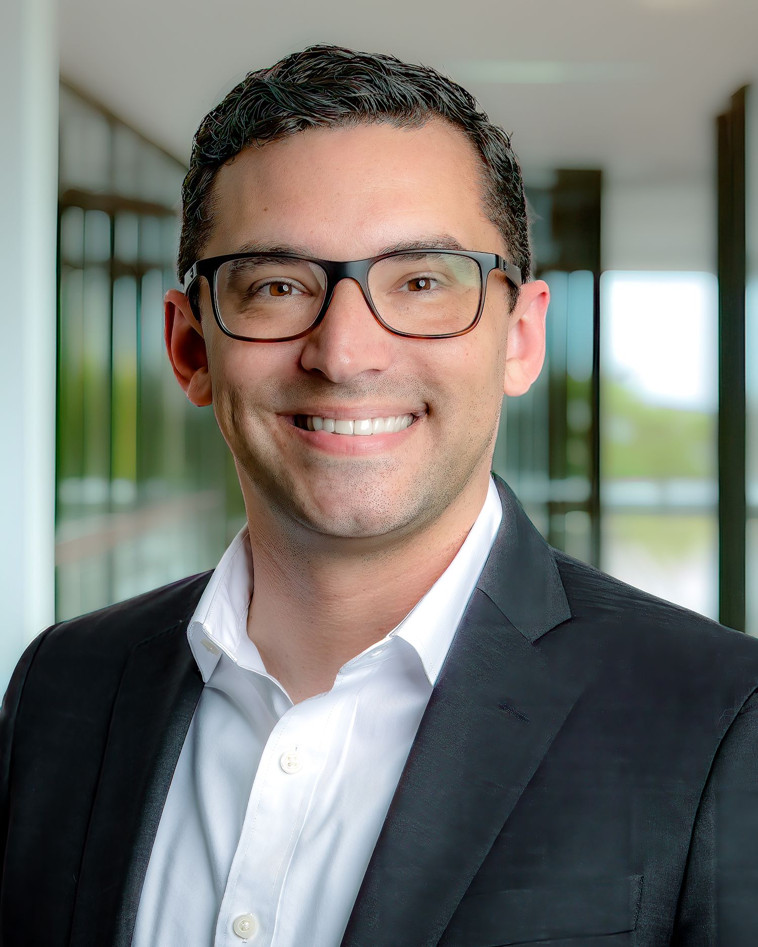 Professional headshot of a smiling man in glasses and a dark blazer with white shirt in a modern office building.
