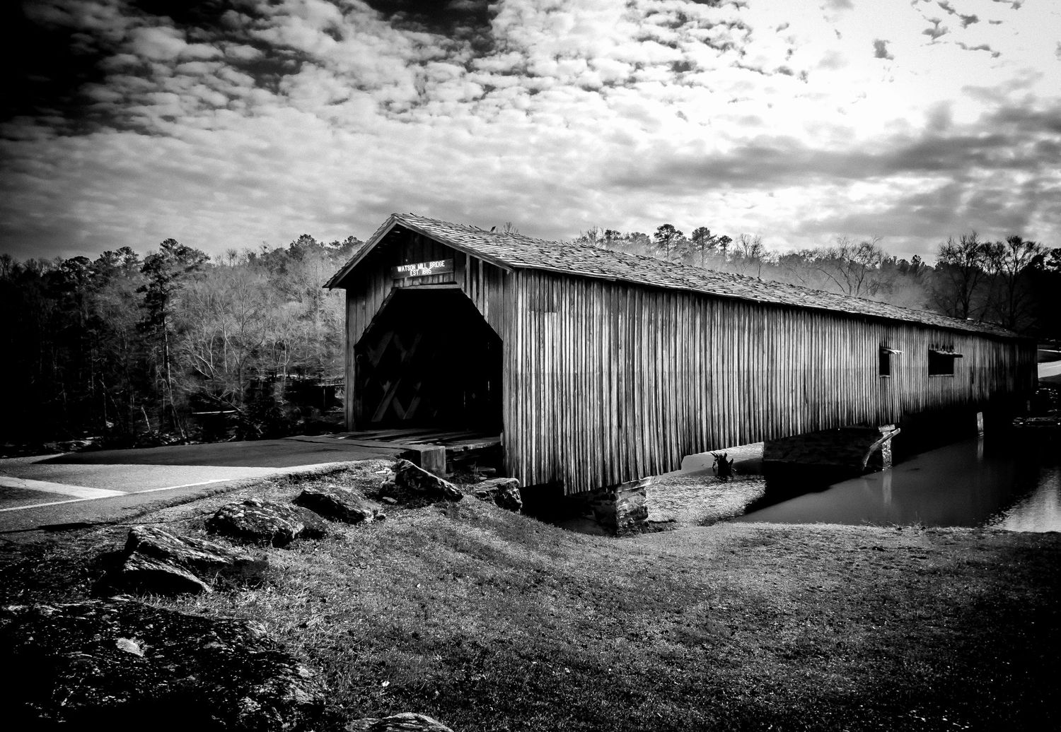 Black and white photo of a rustic wooden covered bridge beside a calm river in a forest.