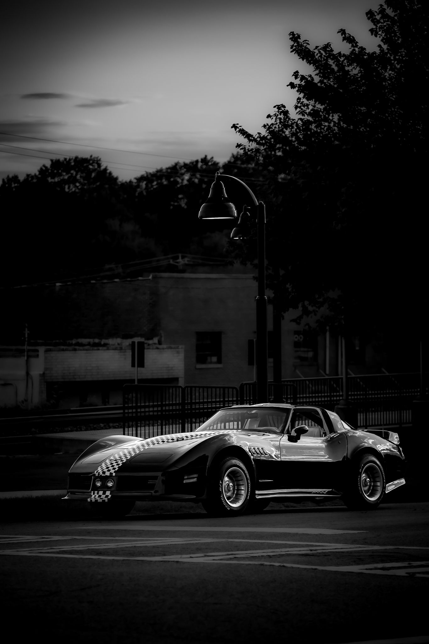 Black and white photo of a classic Corvette sports car with checkered stripe parked on a street at dusk.