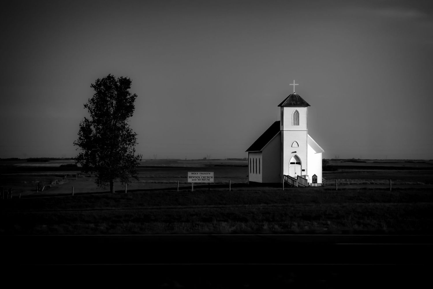Black and white photo of a small white church with a steeple standing alone on a flat open prairie landscape.