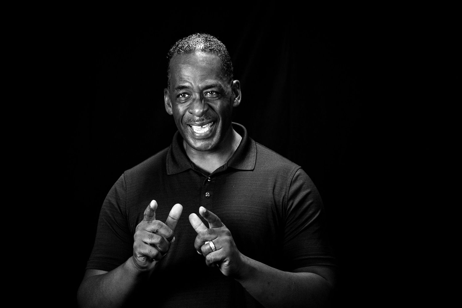 Black and white portrait of a smiling man in a polo shirt gesturing with his hands against a dark background.