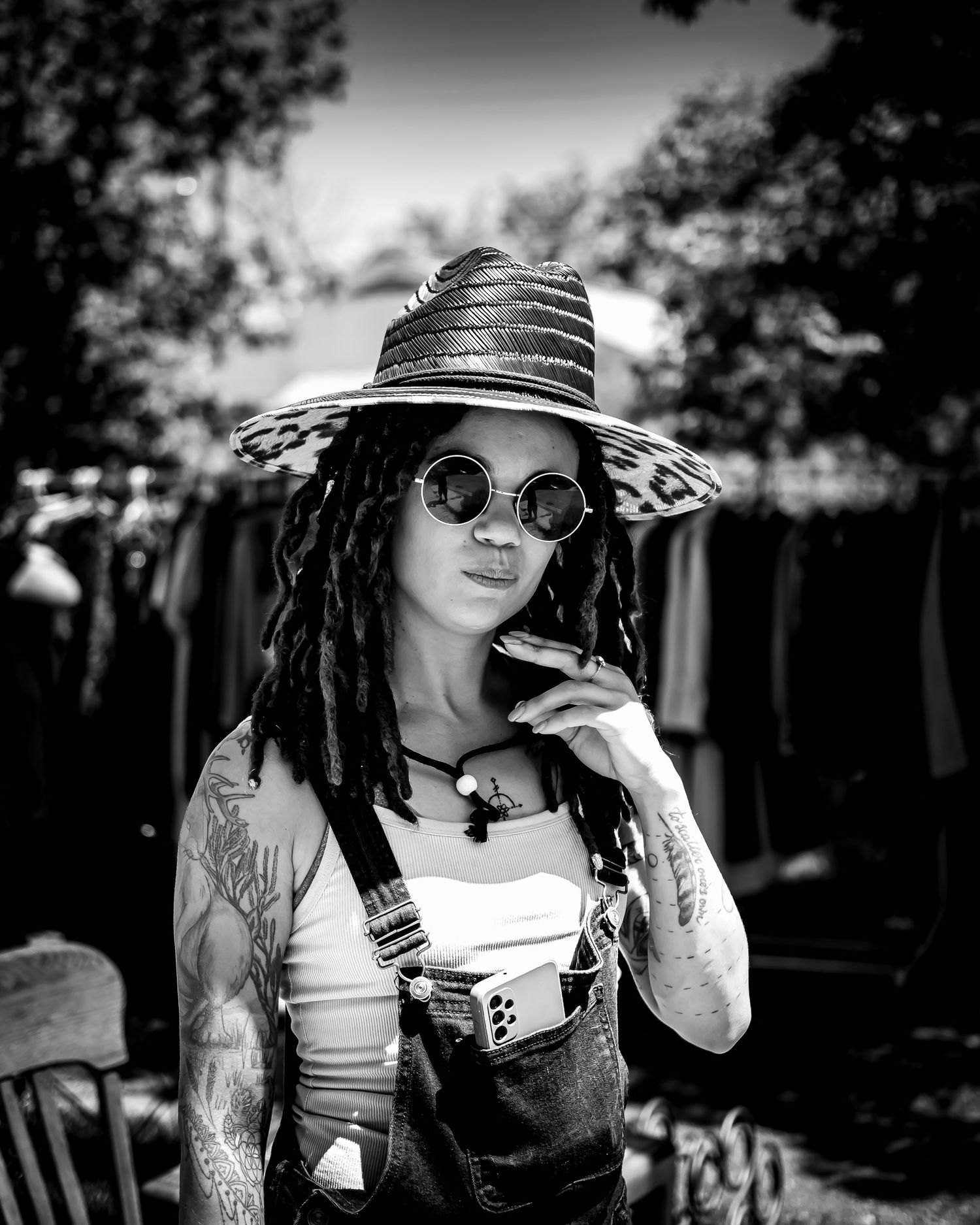 Black and white portrait of a tattooed woman with dreadlocks wearing round sunglasses and a wide-brim straw hat outdoors.