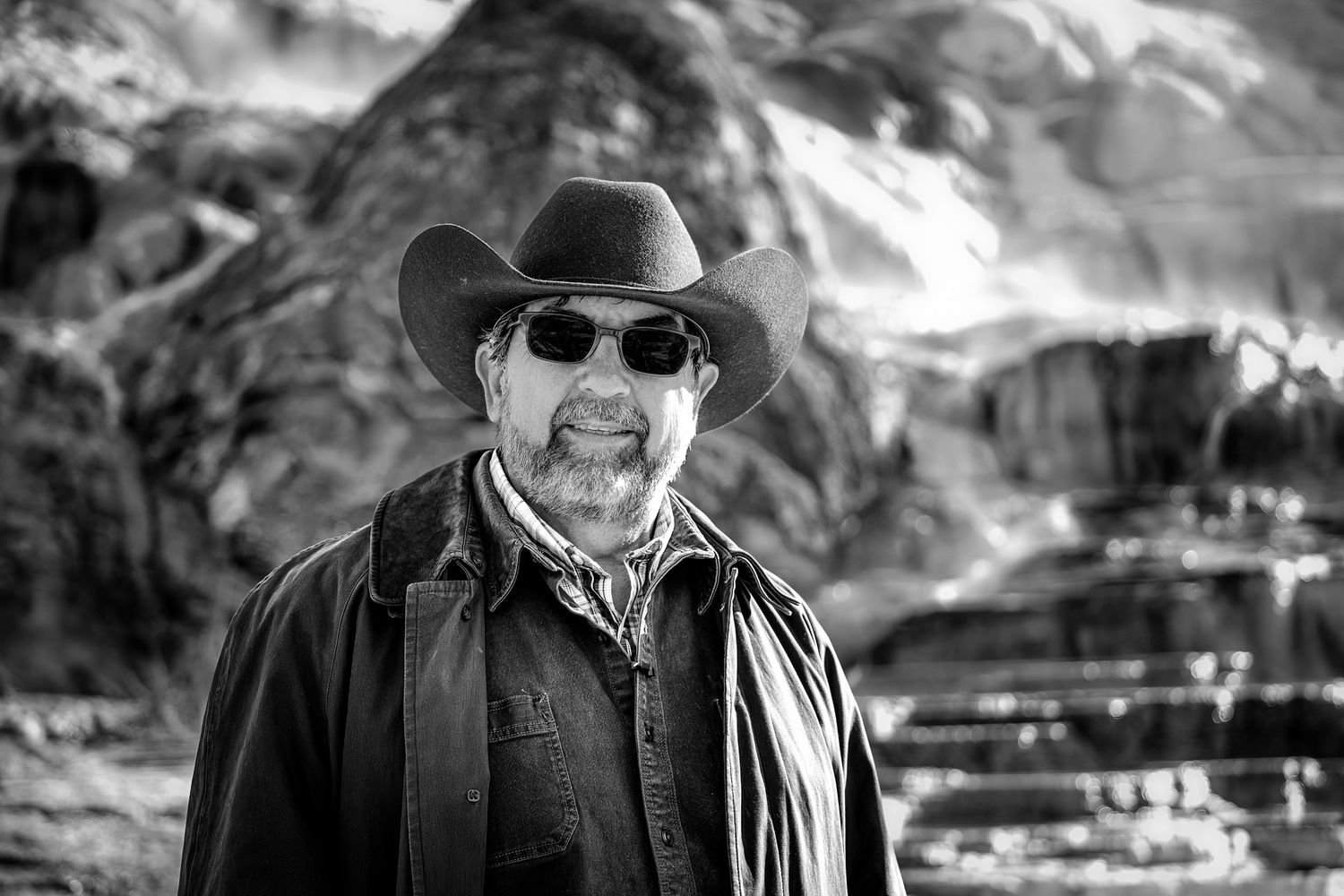 Rugged man in cowboy hat and sunglasses poses before rocky mountain landscape in black and white.