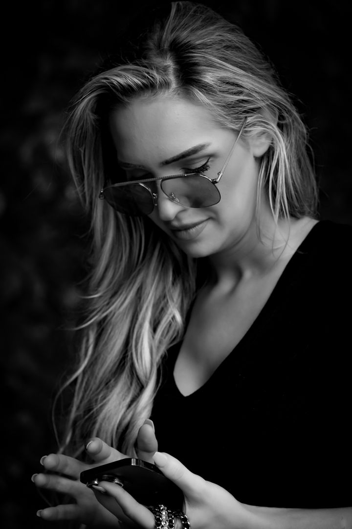 Blonde woman wearing aviator sunglasses looks down at her phone in a black and white portrait.