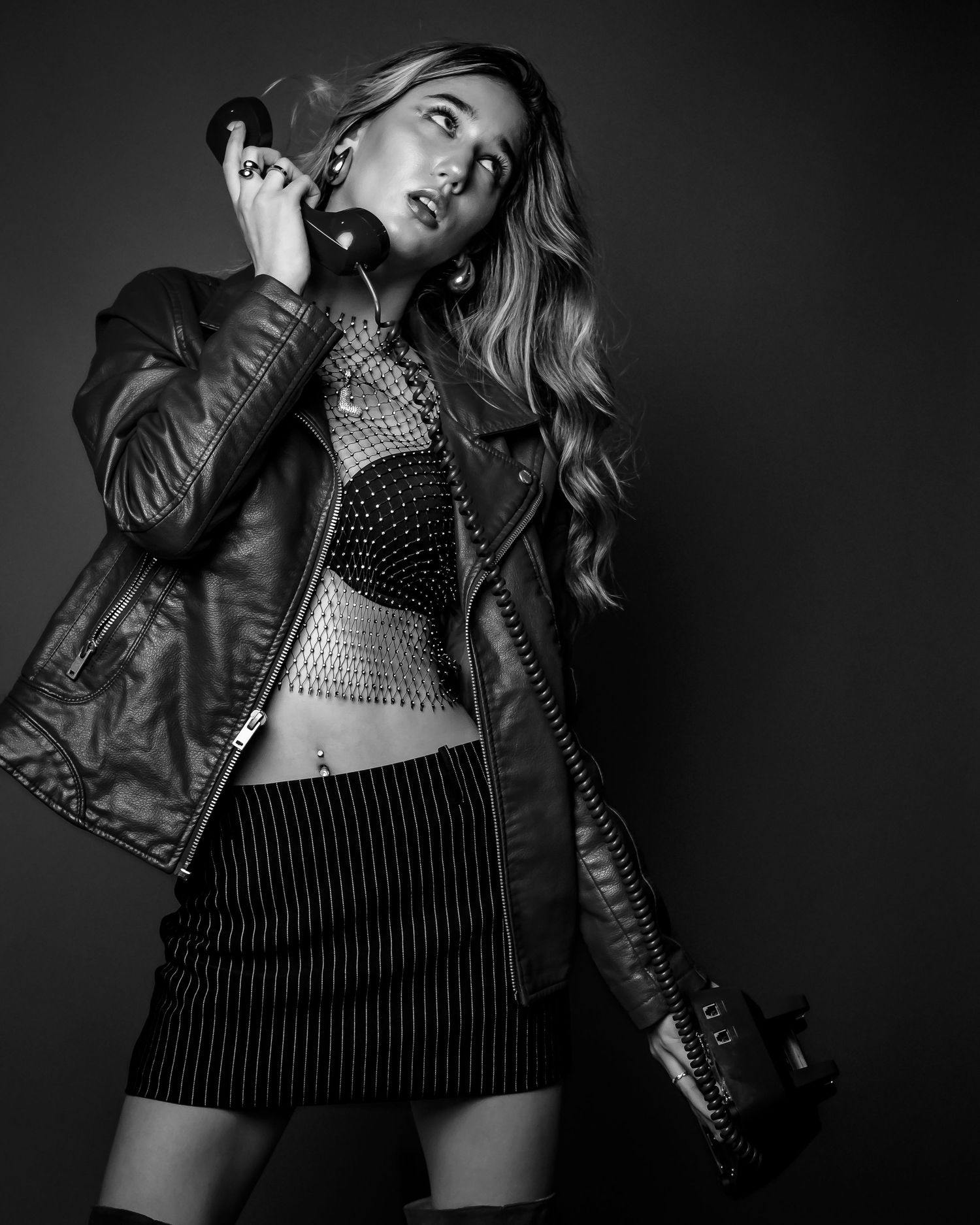 Blonde woman in leather jacket holds vintage phone in dramatic black and white fashion portrait.