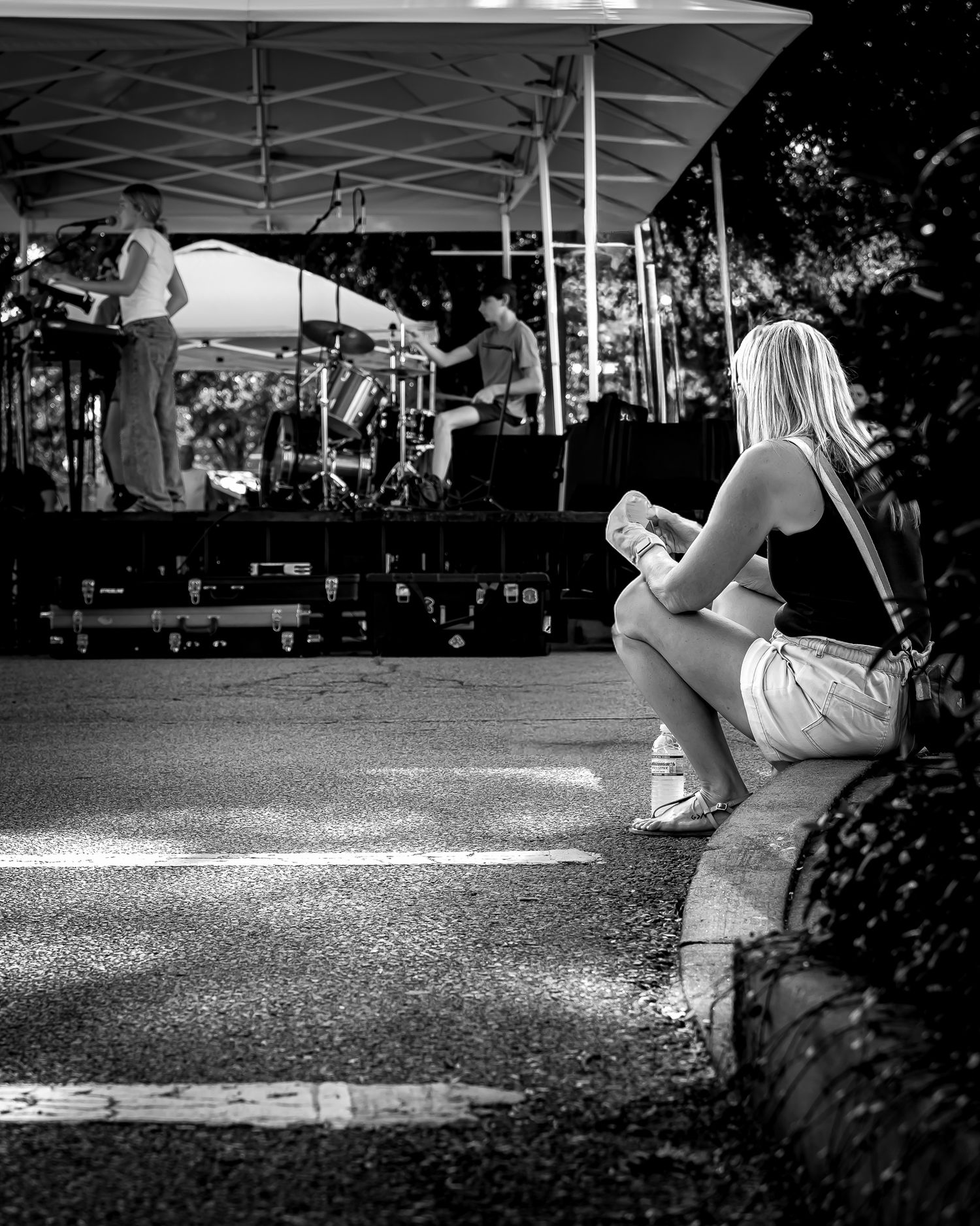 Black and white photo of a blonde woman sitting on a curb watching an outdoor concert stage with musicians performing.