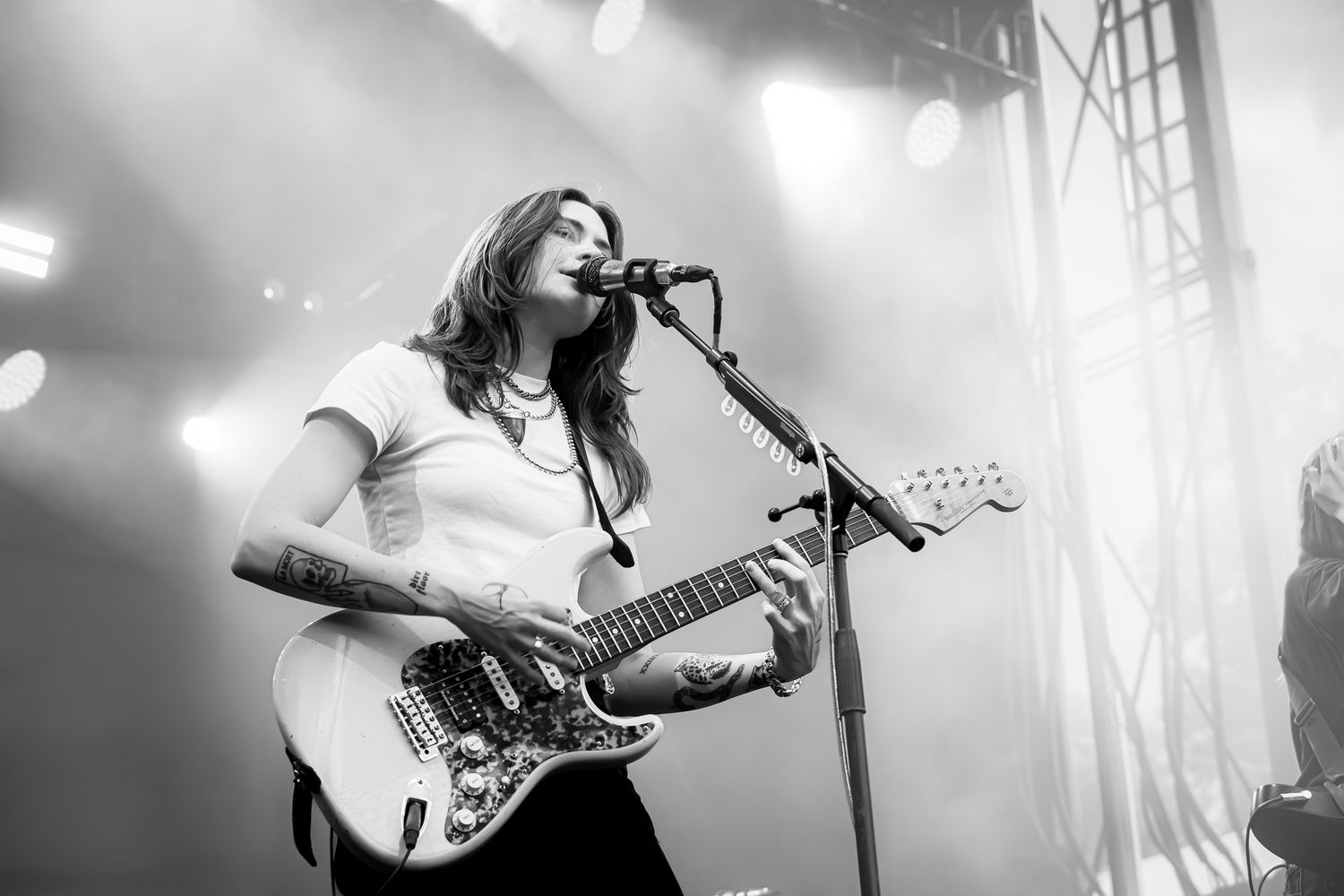Female tattooed guitarist singing into microphone while playing electric guitar on stage in black and white.