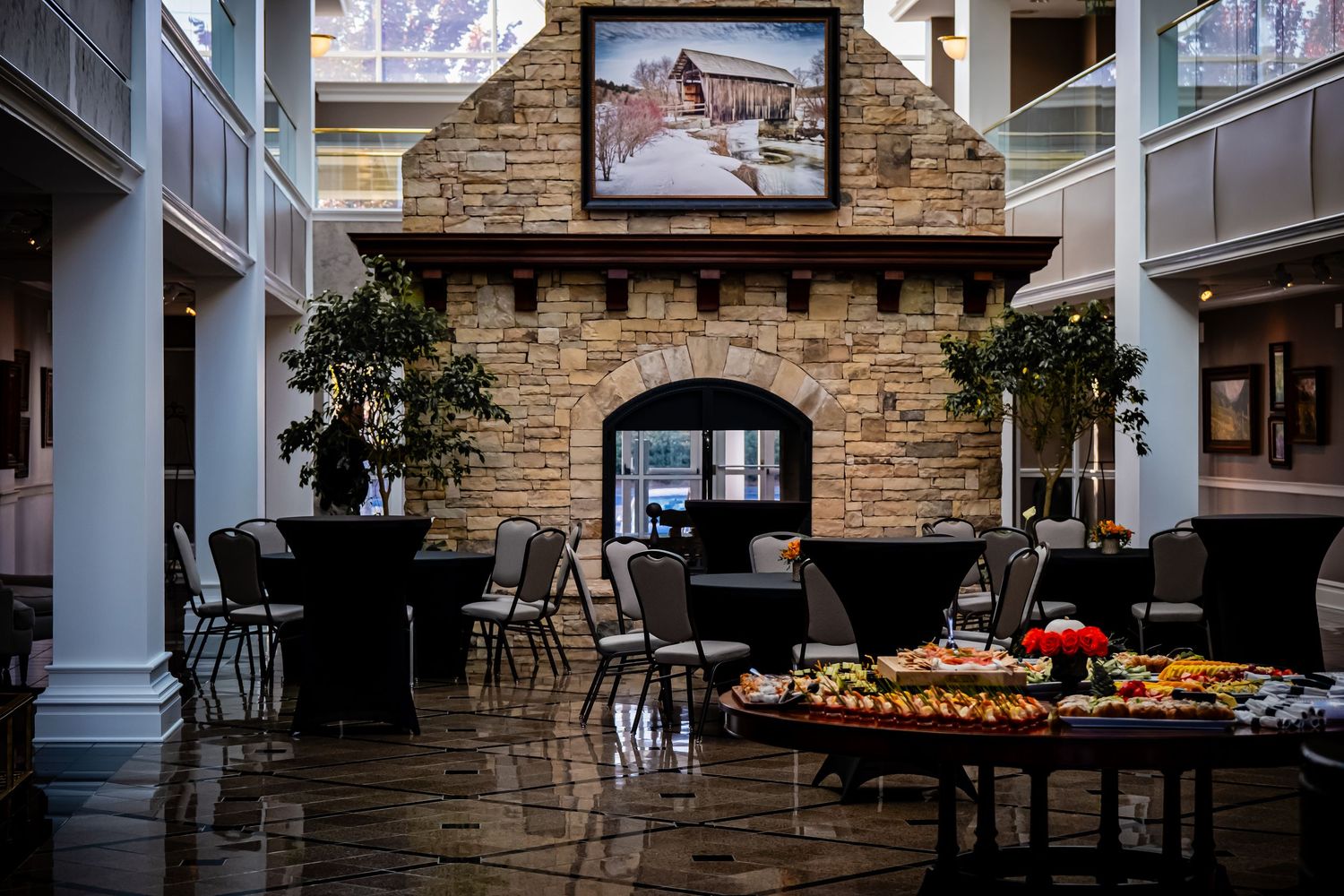 Elegant hotel atrium with stone fireplace, black cocktail tables, and a catered fruit display on a round table.