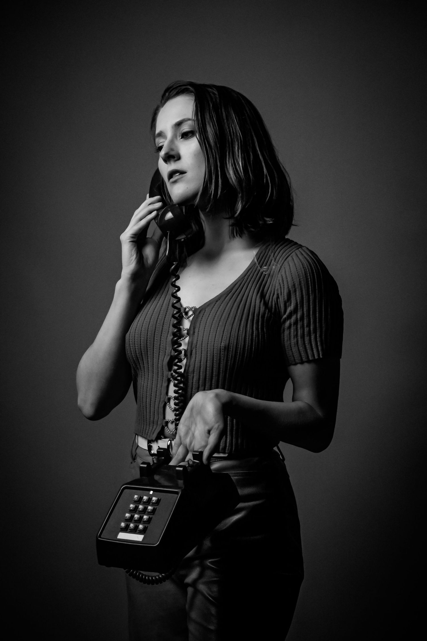 Brunette woman holding vintage rotary telephone poses in moody black and white studio portrait.