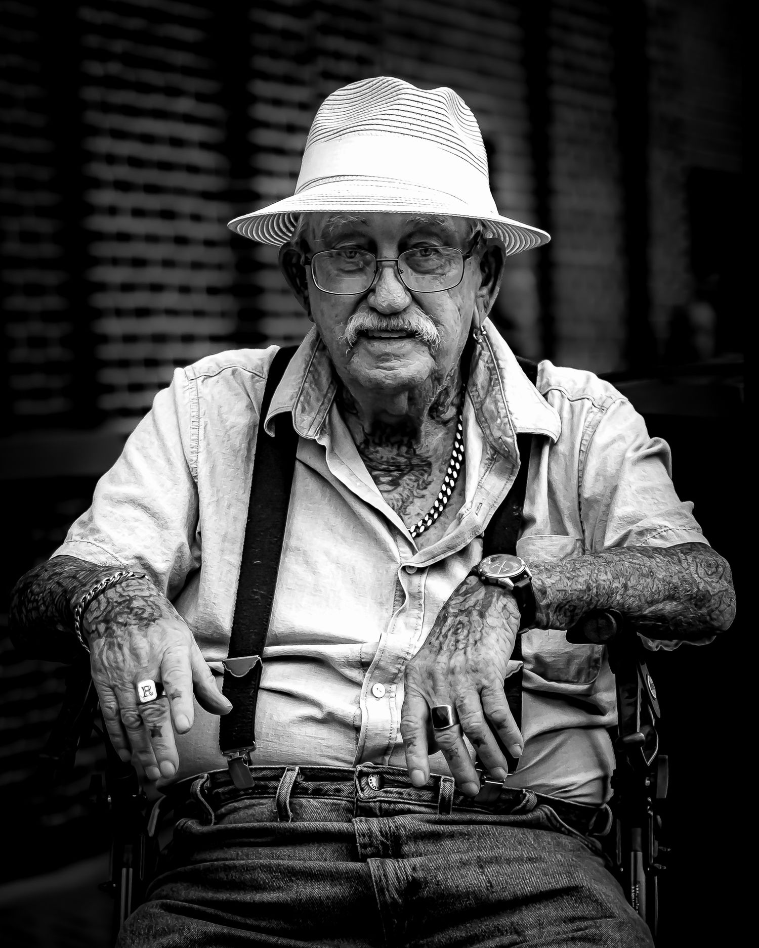 Elderly tattooed man in white fedora, glasses, and suspenders sits confidently in black and white portrait photo.
