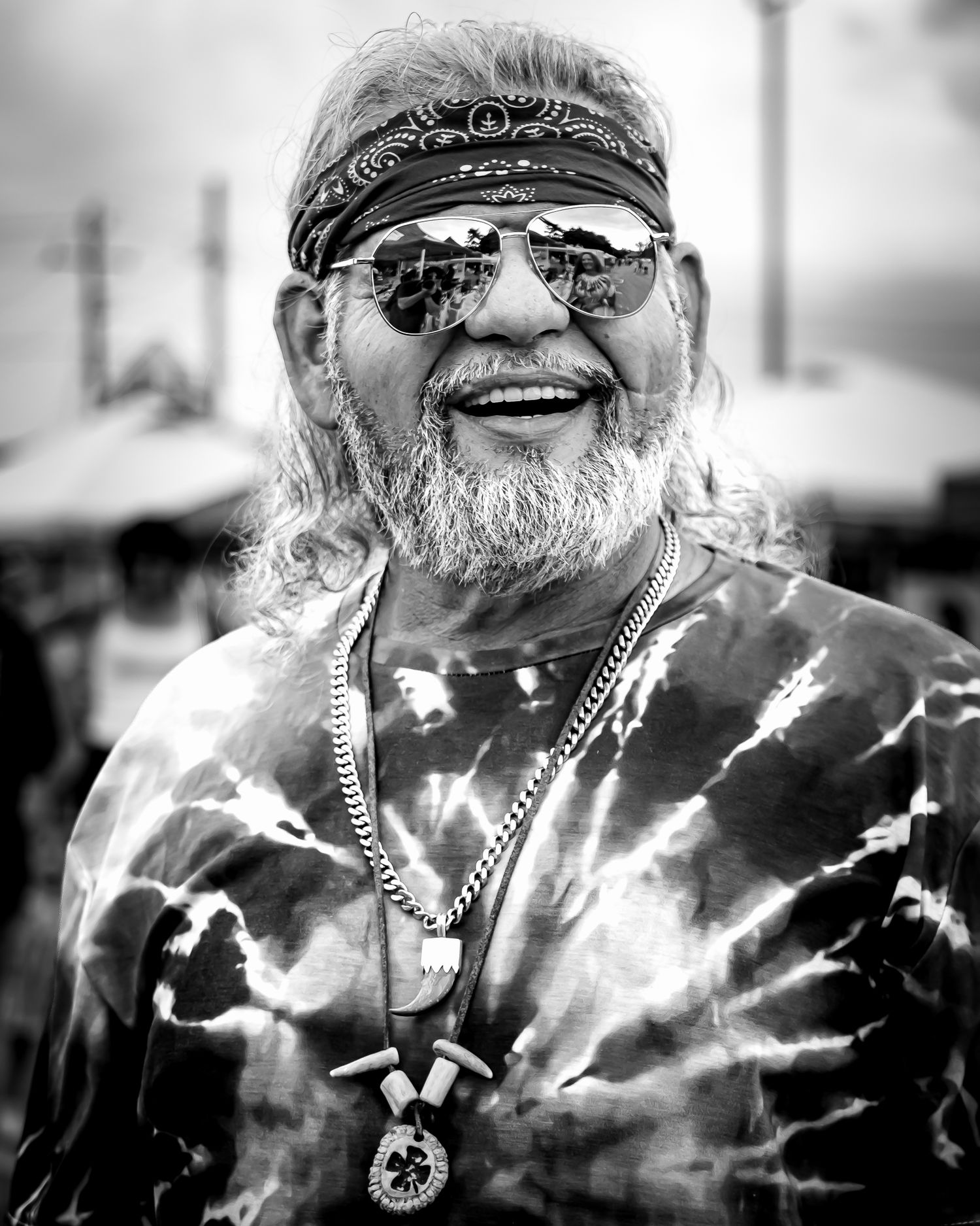 Smiling older man with gray beard, bandana, sunglasses, and tie-dye shirt wearing chain necklaces in black and white photo.