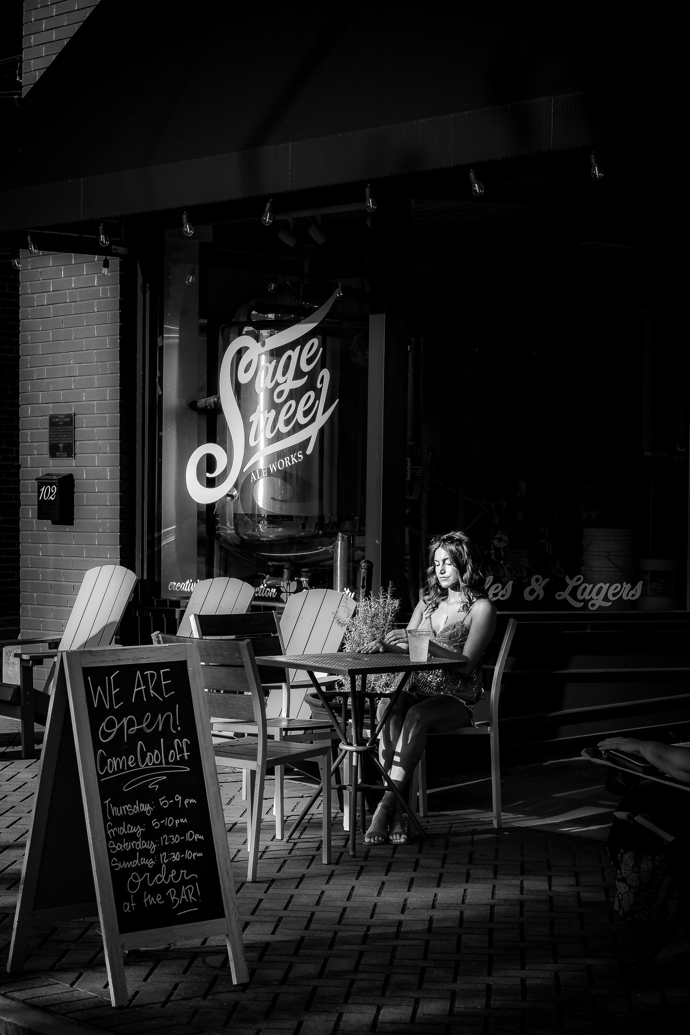 Black and white photo of a woman sitting alone outside Sage Steel Works brewery at night, with an open sign on the sidewalk.