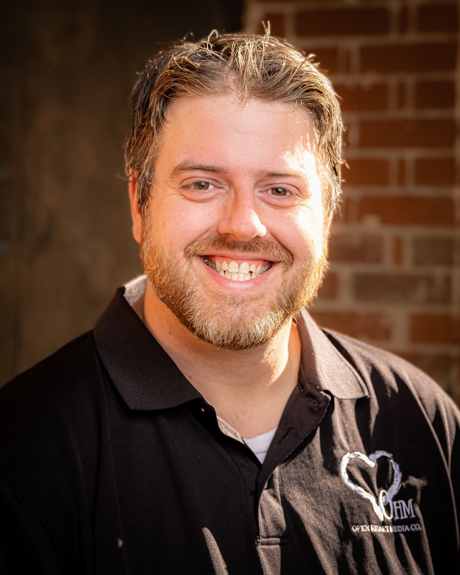 Smiling bearded man in black polo shirt posing outdoors against a rustic brick wall background.