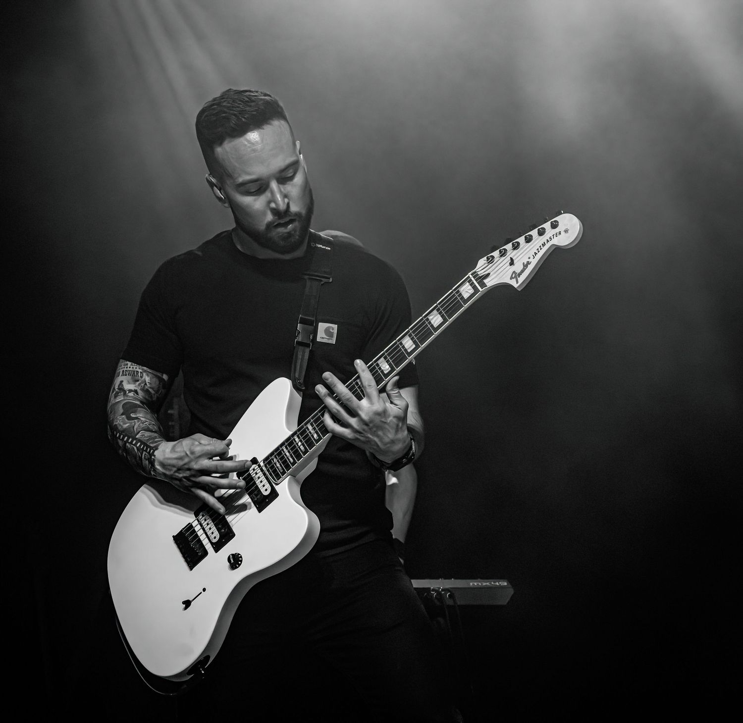 Black and white photo of a tattooed guitarist playing a white electric guitar on a smoky concert stage.