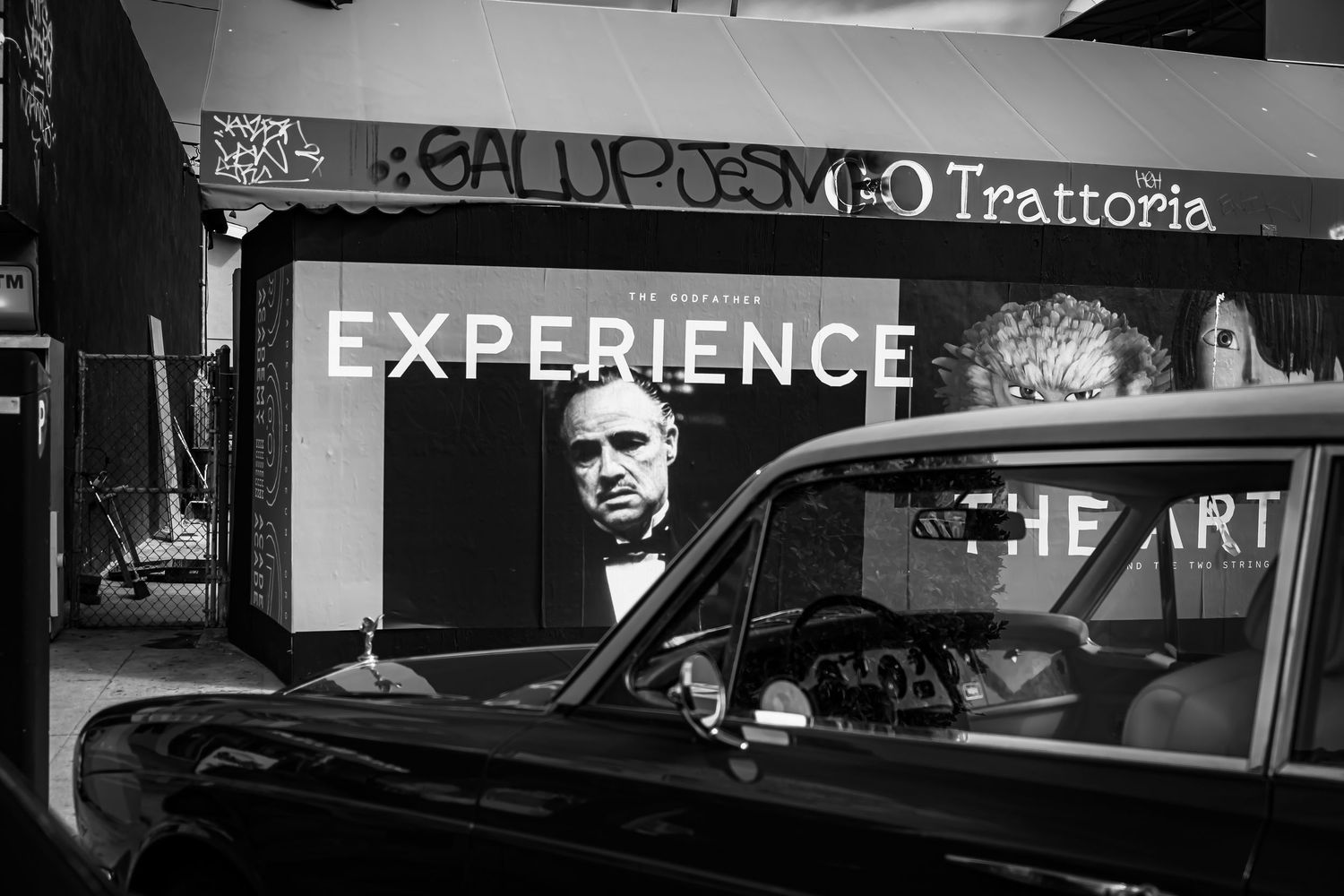 Classic black vintage car parked outside Trattoria restaurant with Godfather Experience billboard in black and white.