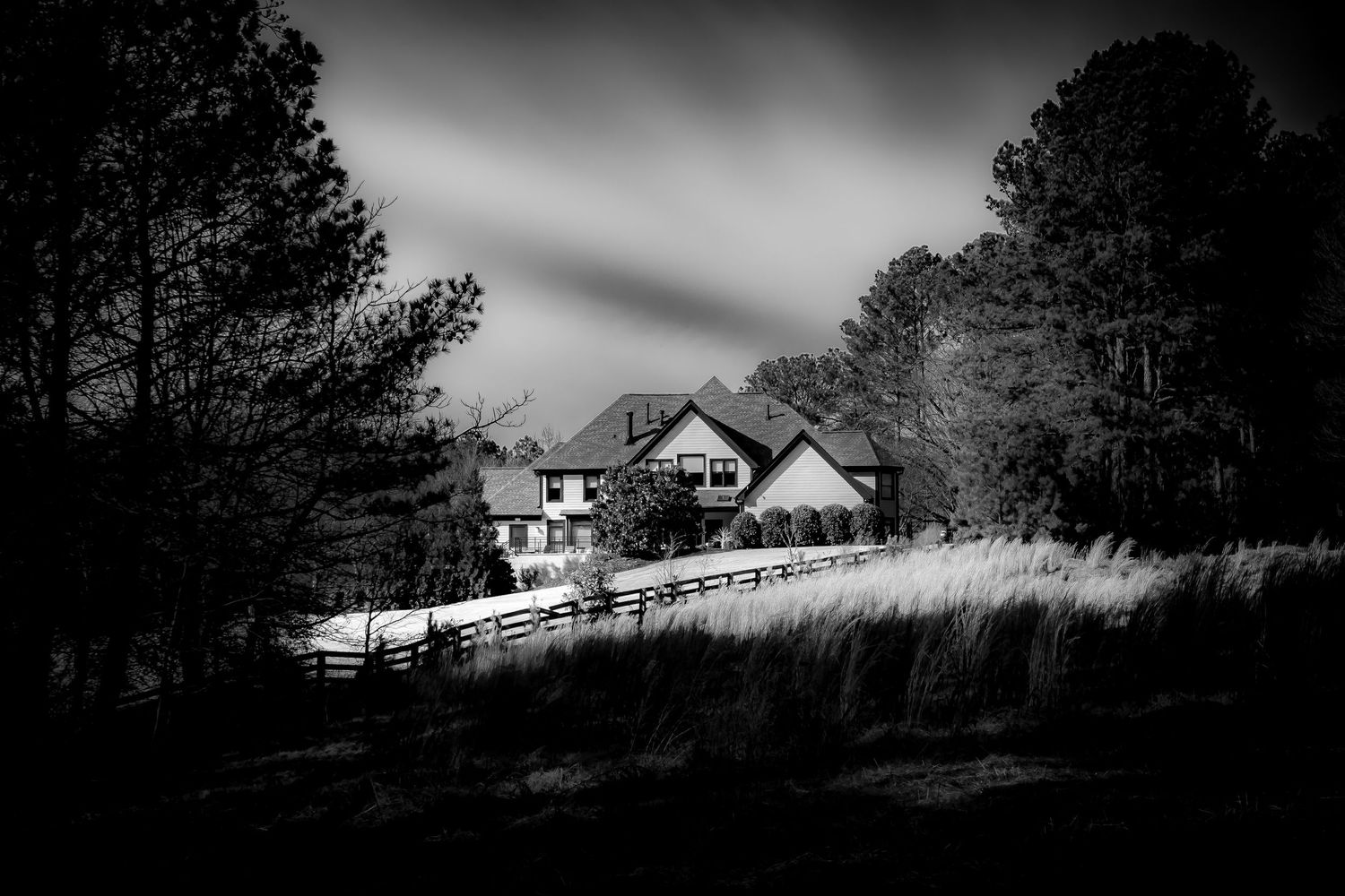 Black and white photo of a large house surrounded by trees and a wooden fence under moody skies.