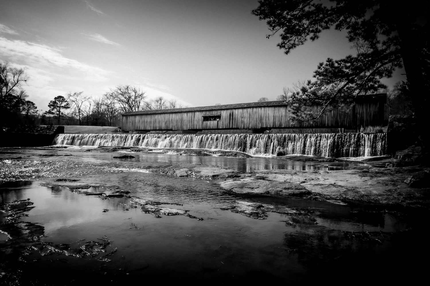 Black and white photo of a historic covered bridge spanning a cascading waterfall over rocks.