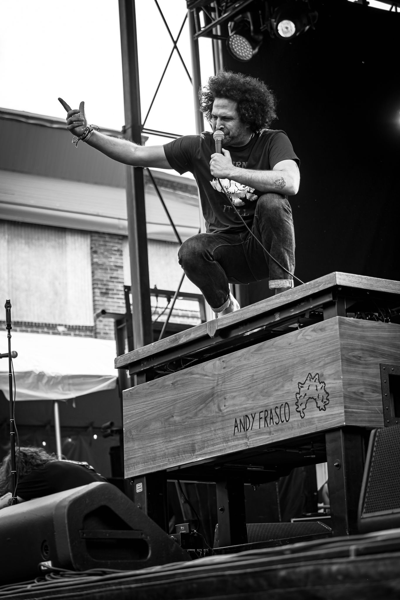 Andy Frasco crouching on wooden speaker box gesturing to crowd while holding microphone on stage.