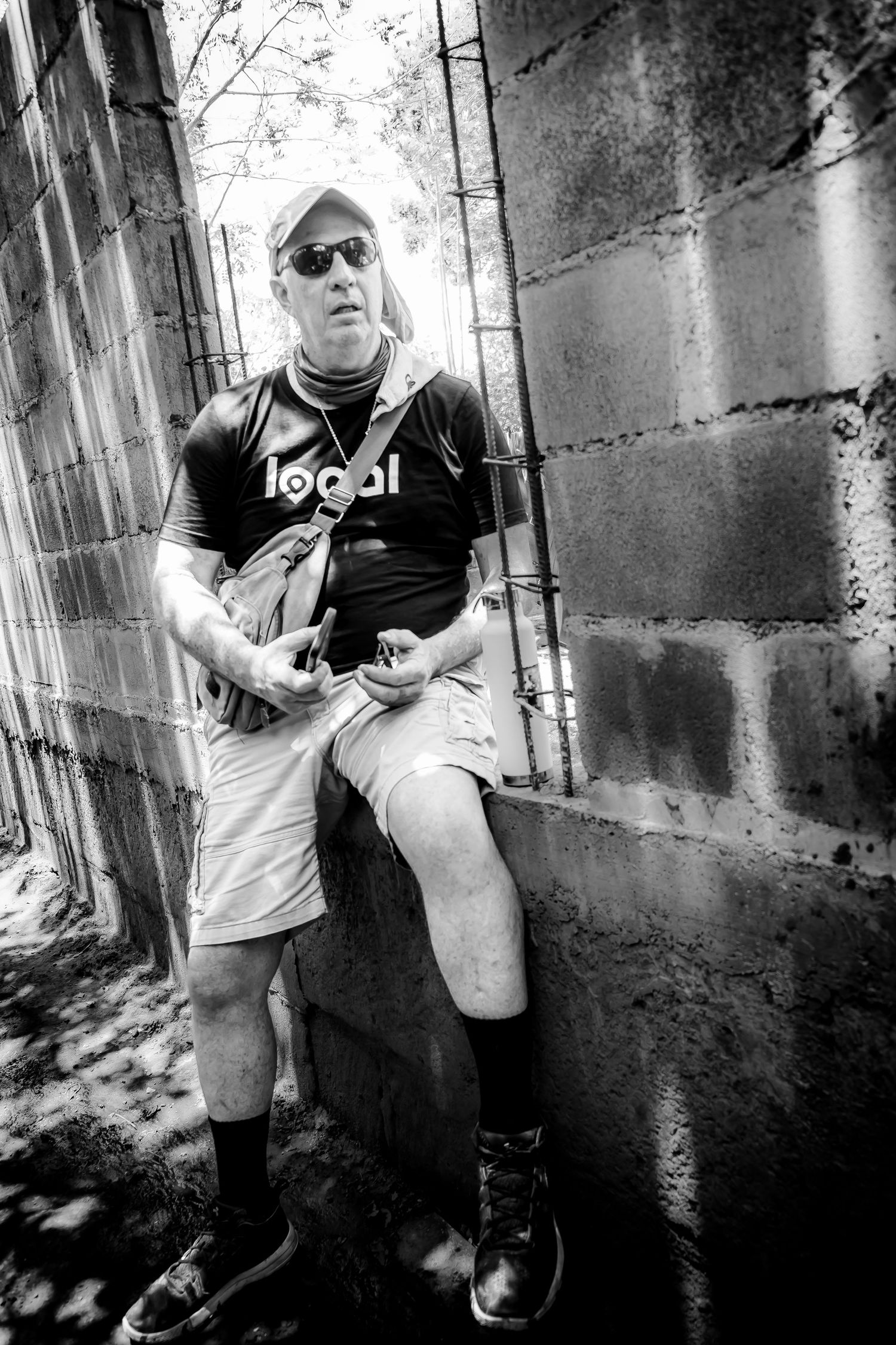 Black and white photo of a man in sunglasses and a Local t-shirt sitting against a concrete block wall outdoors.