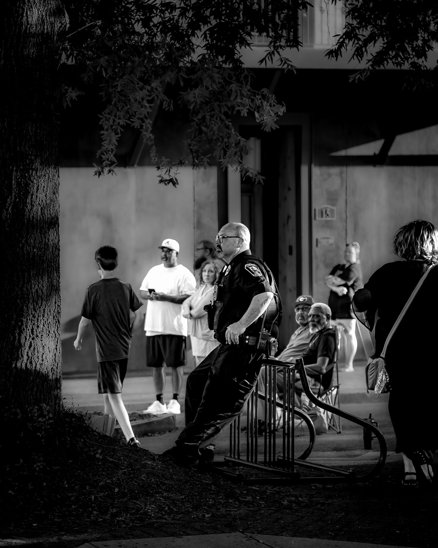 Black and white photo of a police officer standing near a railing outside a building with pedestrians nearby.