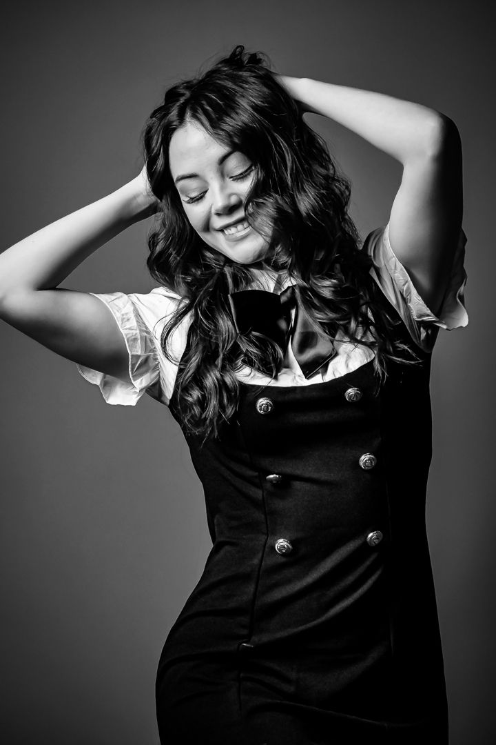 Black and white photo of a smiling young woman in a dark pinafore dress with hands raised in her wavy hair.