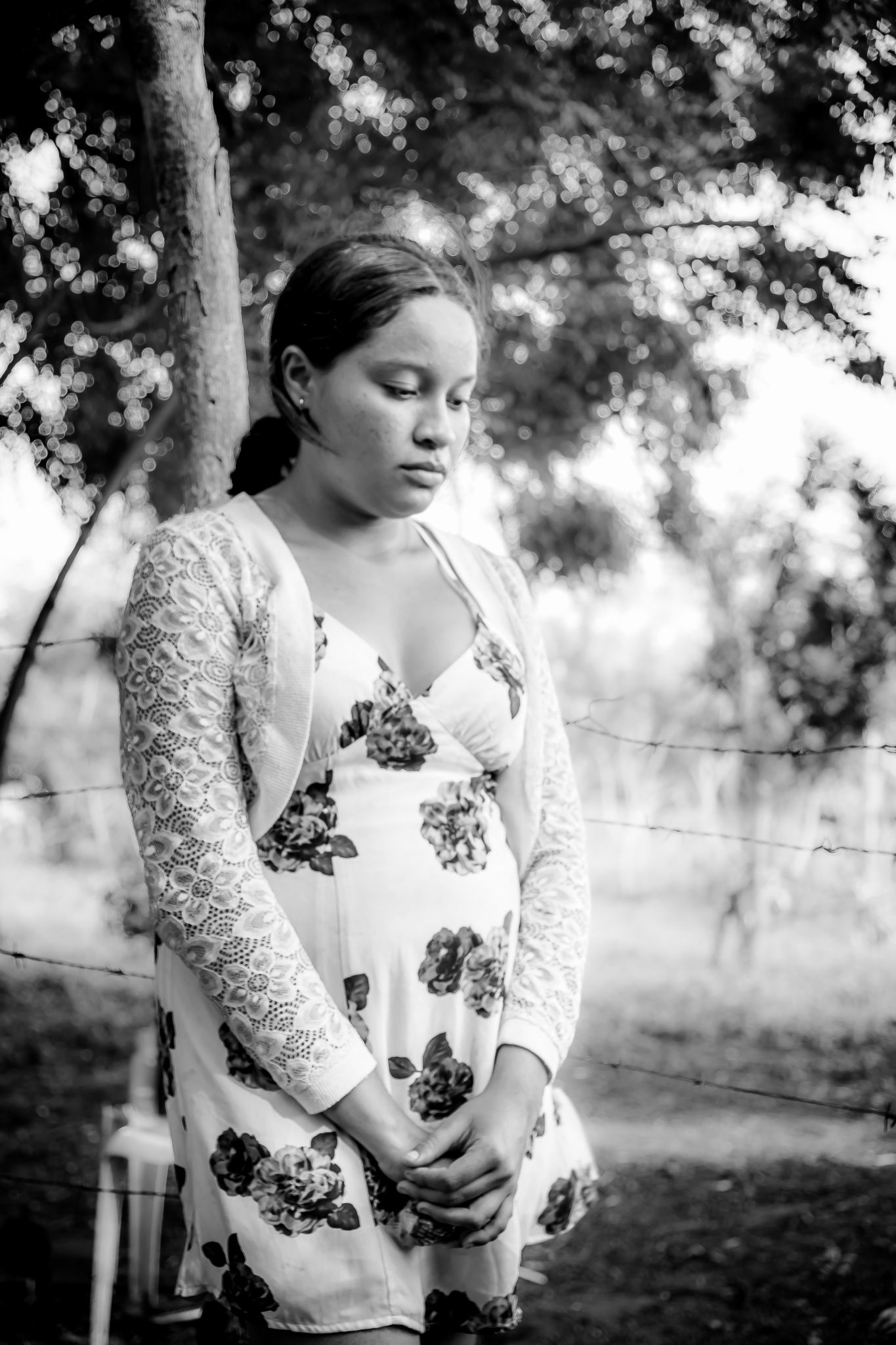 Young pregnant woman in floral dress standing outdoors near trees in black and white.