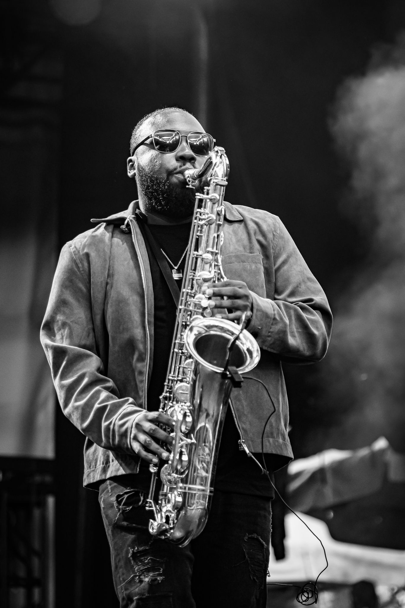 Bearded male saxophonist wearing sunglasses performing on stage in black and white photo.