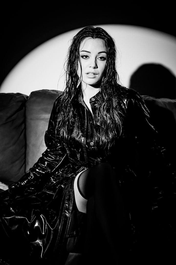 Black and white photo of a woman with wet hair in a patent leather coat seated on a sofa with circular backdrop.