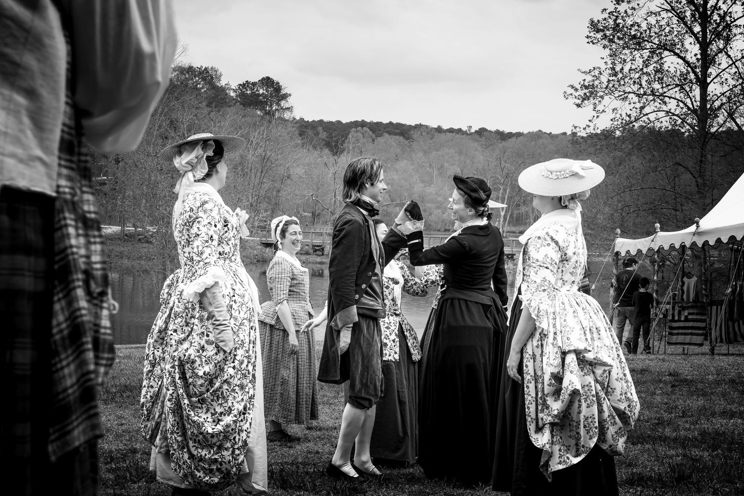 Group of women in 18th century colonial dresses gather on open field, black and white reenactment scene.