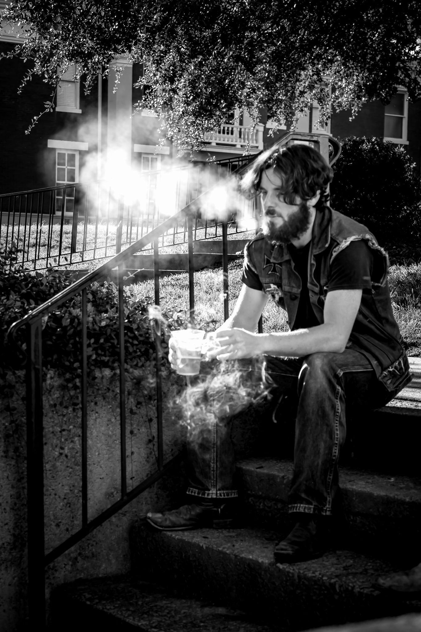 Black and white photo of bearded man sitting on steps holding smoking container outdoors near iron railing.