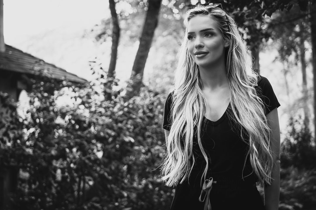 Young blonde woman with long hair stands outdoors among trees in a black and white portrait.