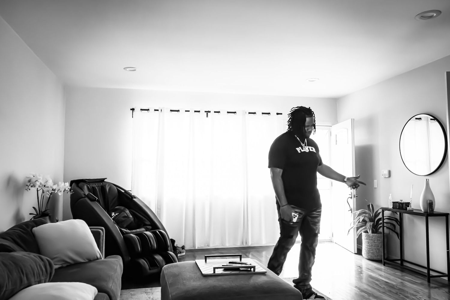 Black and white photo of a man standing in a modern living room with large curtained windows and hardwood floors.