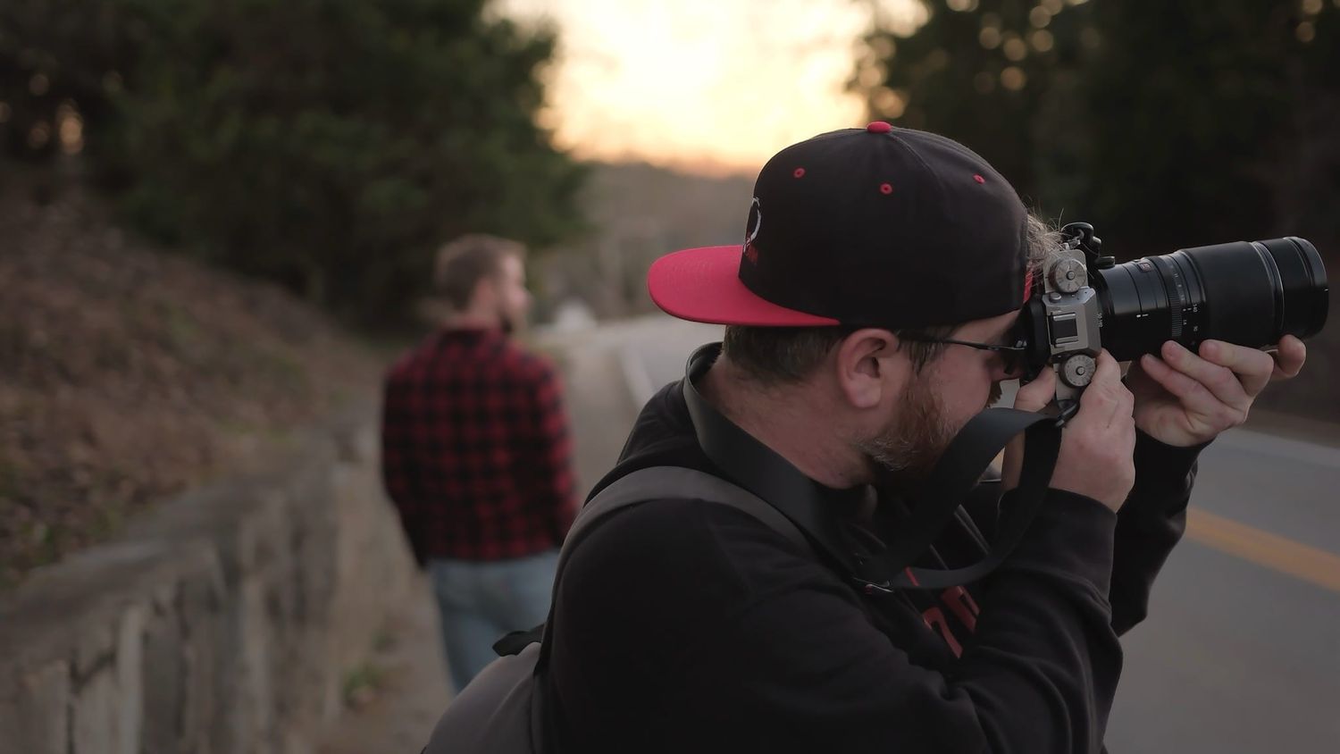 Photographer with a red and black cap uses a telephoto lens to capture a subject on a tree-lined road at sunset.
