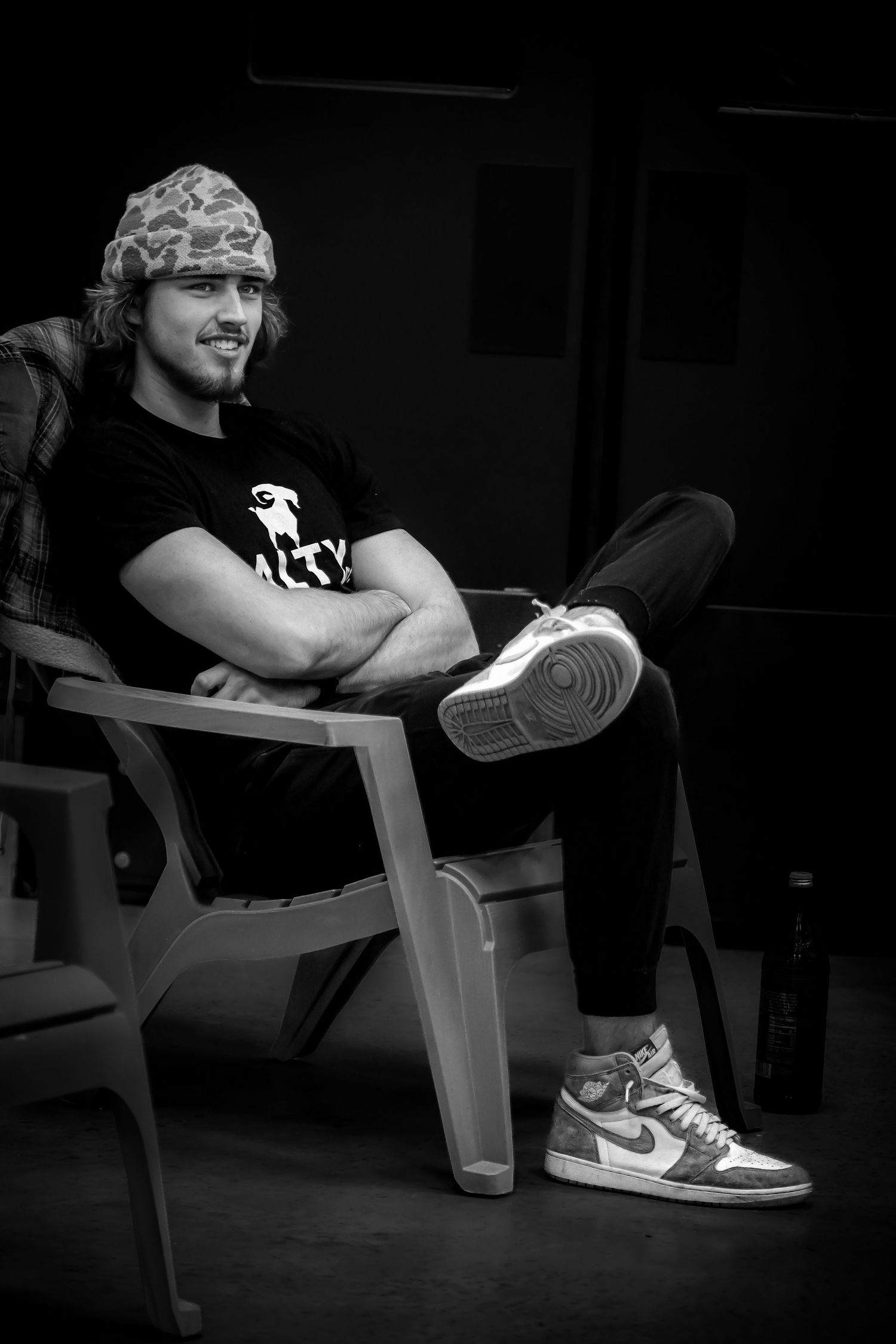 Smiling man in camo hat and Nike sneakers relaxes in chair in black and white portrait. Podcast coverage. Branding. Branding material.