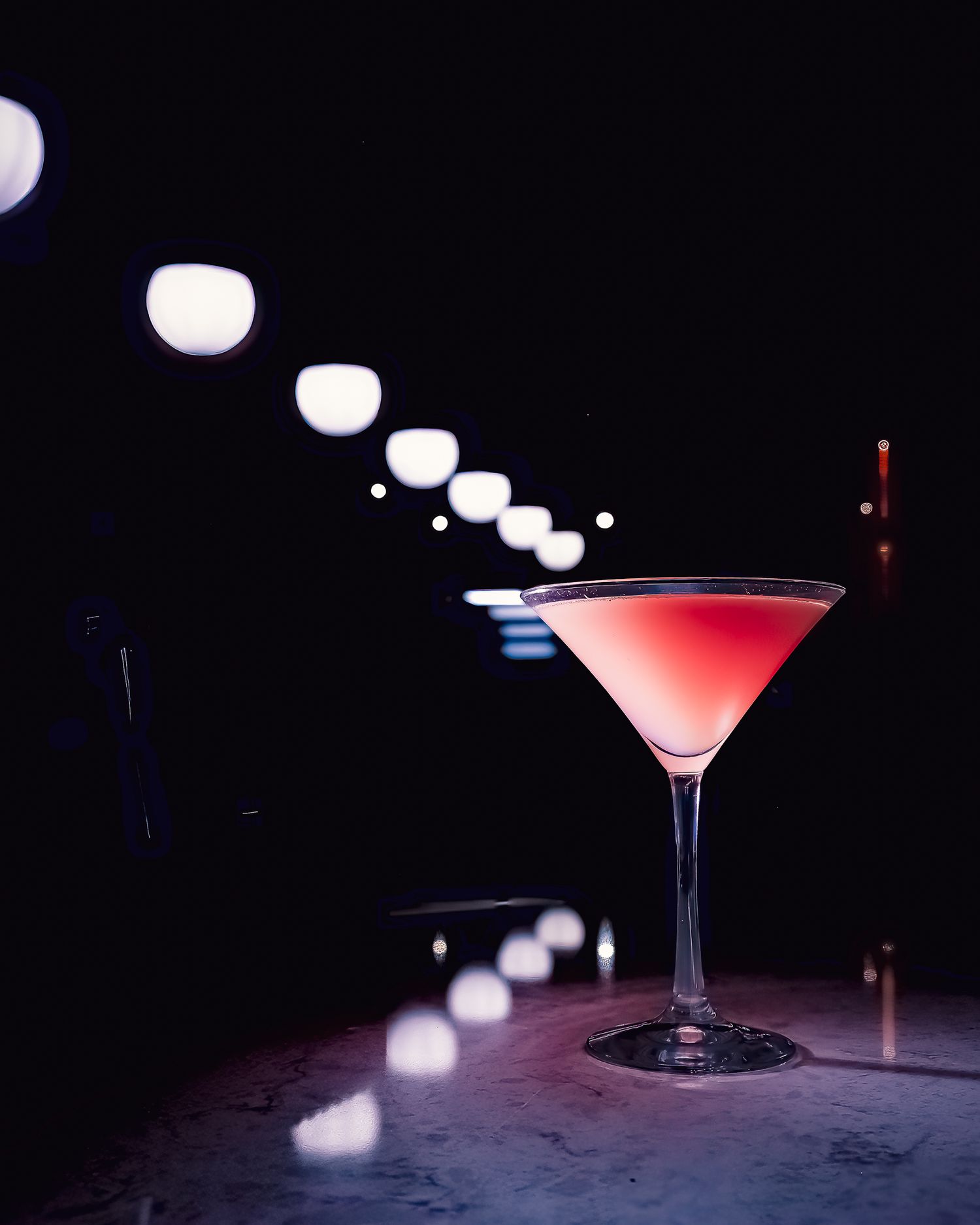 A pink cocktail in a martini glass on a dark bar counter with bokeh lights in the background. Product photography. Bar photography. Branding