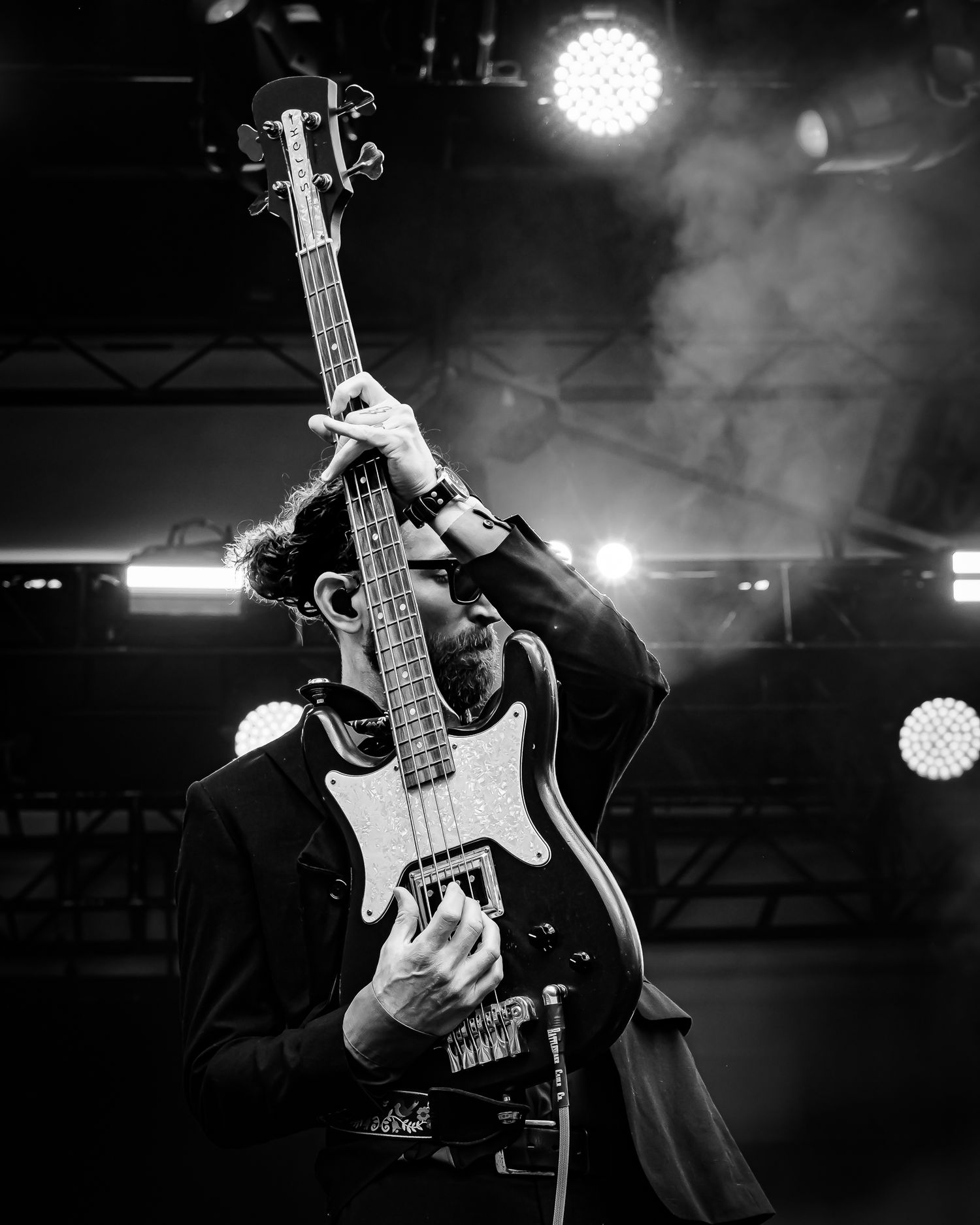 Black and white photo of a bearded male bassist raising his guitar overhead dramatically on a smoky stage.