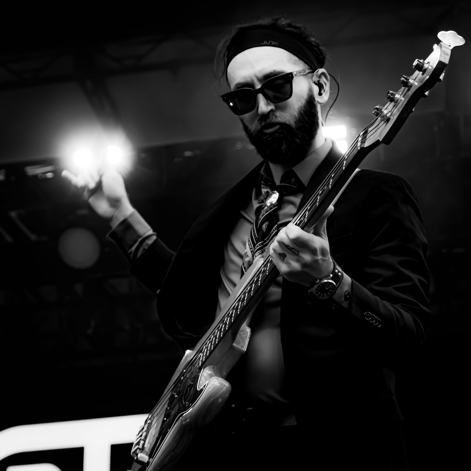 Black and white photo of a bearded male bassist in a suit and sunglasses performing under stage lights.