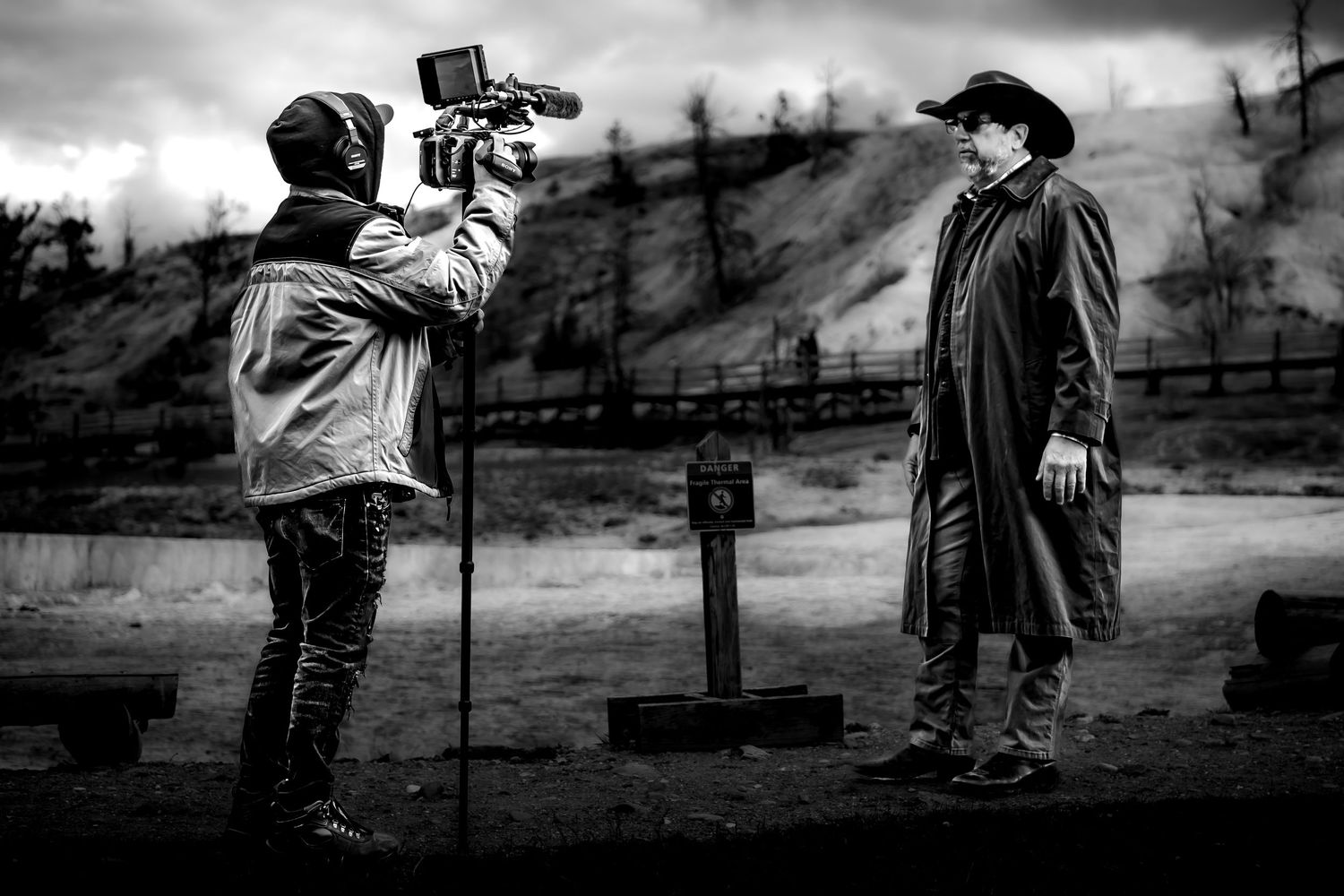 Black and white behind-the-scenes film shoot with cameraman filming actor in cowboy hat and long coat outdoors.